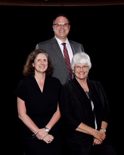 Formal studio portrait of three smiling adults against a black background. Two women sit in front while a man in a charcoal suit and red patterned tie stands behind them.
