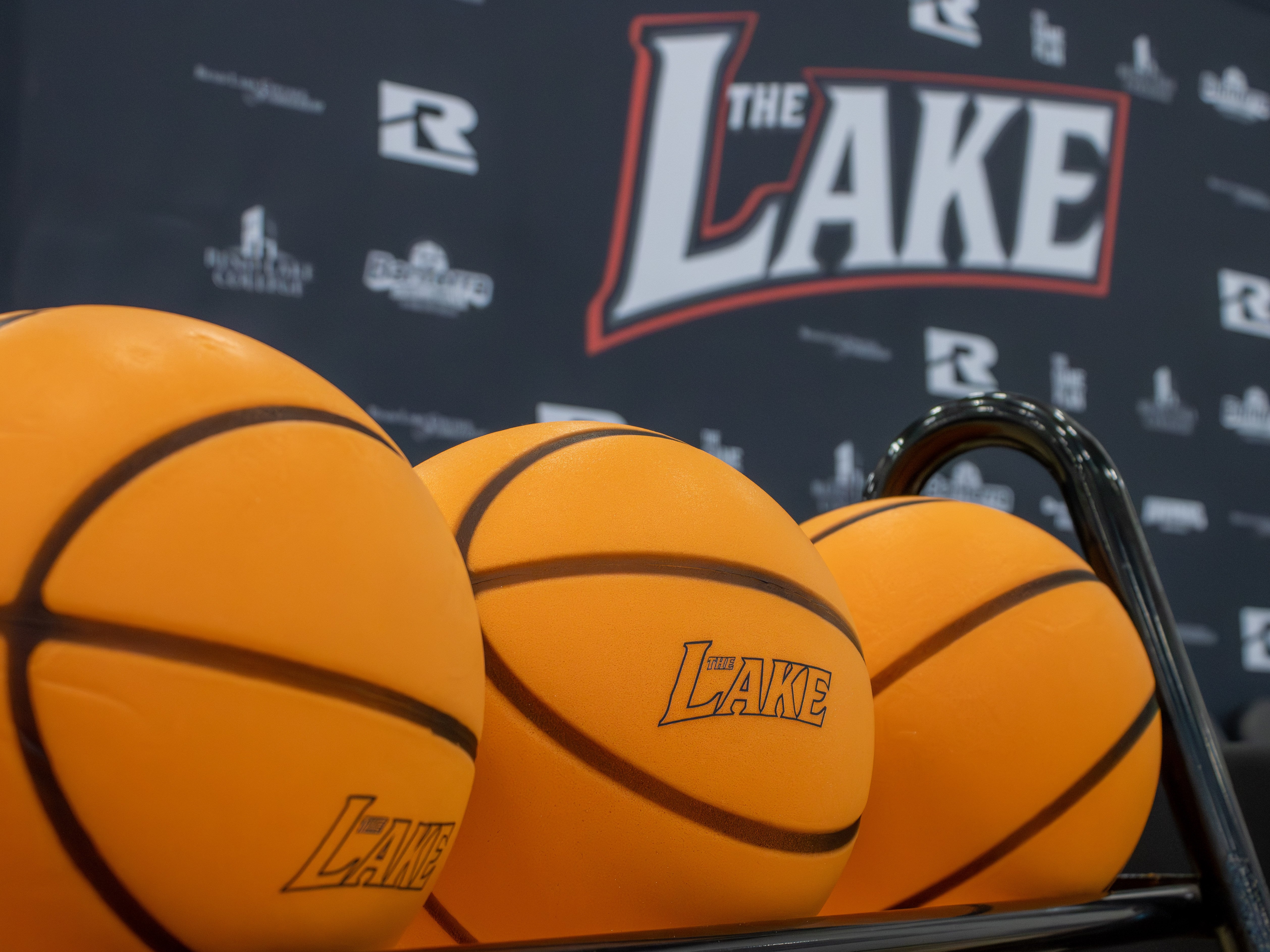 A close-up shot of three orange basketballs resting on a rack, each featuring "The Lake" logo. In the background, a blurred black backdrop displays logos for Rend Lake College, Banterra Sports Complex, and "The Game Plan".