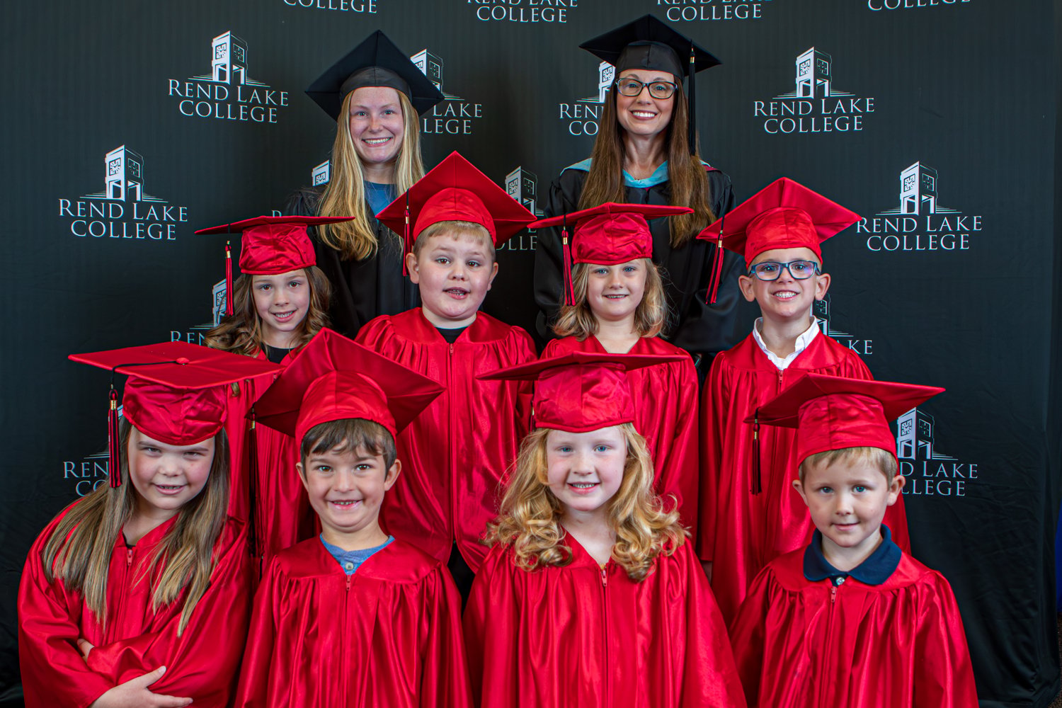 A group graduation portrait featuring eight young Pre-K graduates and two teachers posed against a black step-and-repeat backdrop covered in white "Rend Lake College" logos. The students are arranged in two rows wearing shiny red graduation gowns and matching red mortarboard caps.