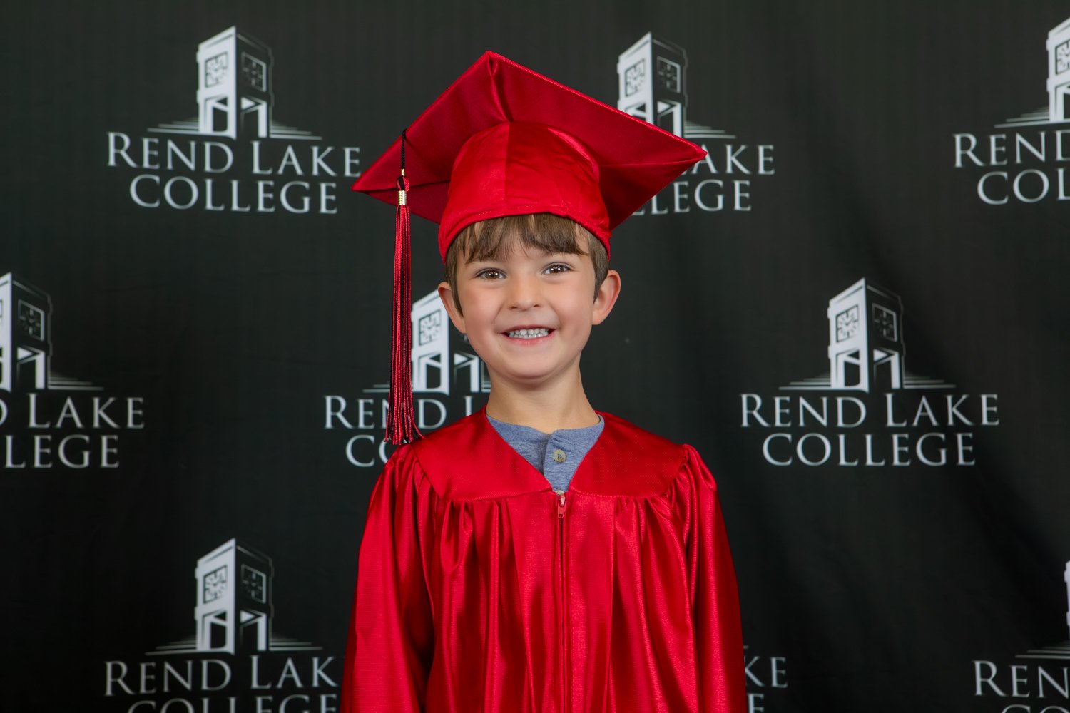 A studio portrait of a young boy with light brown hair and front bangs showing a wide, toothy smile. He is dressed in a vibrant red graduation gown over a grey button-up collar, topped with a matching red mortarboard cap. The backdrop is black, featuring a repeating white pattern of the "Rend Lake College" name and institutional logo.