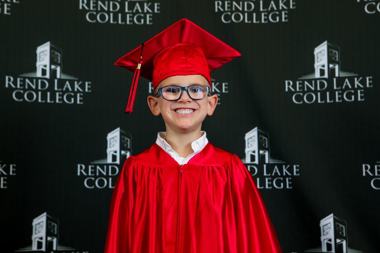 A studio portrait of a young boy with short hair and black-rimmed glasses smiling broadly. He wears a crisp white collared shirt underneath a shiny red graduation gown and a matching red cap. He is positioned in front of a repeating white "Rend Lake College" logo on a solid black background.