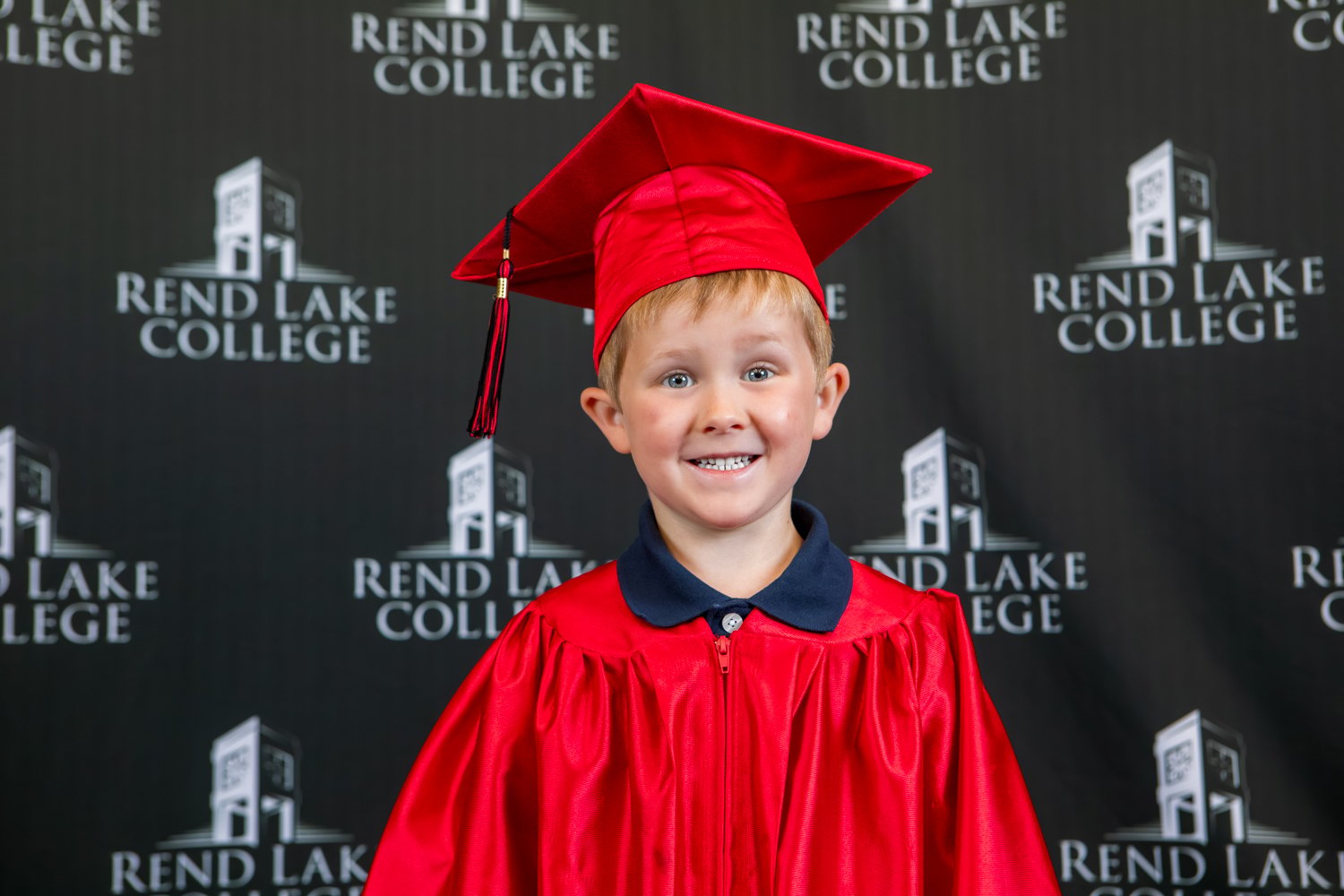 A studio portrait of a young boy with short blonde hair grinning happily. He is wearing a dark blue polo shirt beneath a glossy red graduation gown, paired with a matching red mortarboard cap. The background is a black step-and-repeat wall displaying the "Rend Lake College" logo in white.