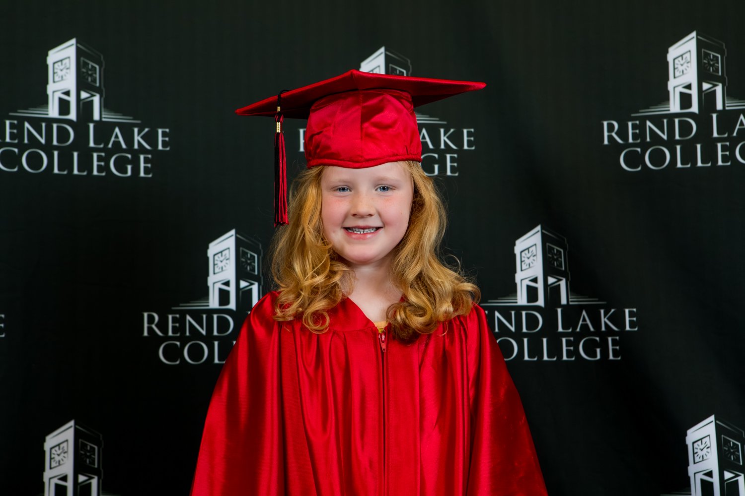 A studio portrait of a young girl with long, wavy blonde hair smiling gently at the camera. She wears a bright red graduation gown and a matching red cap with a tassel hanging on her right side. The background consists of a black studio backdrop with the white "Rend Lake College" logo repeated throughout.