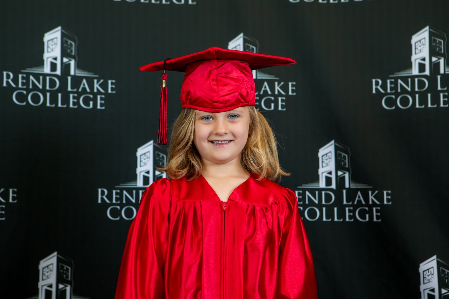 A studio portrait of a young girl with shoulder-length, light brown hair smiling brightly. She is wearing a shiny red graduation gown and a matching red mortarboard cap. She stands centered against a black backdrop covered in a repeating white "Rend Lake College" logo pattern.