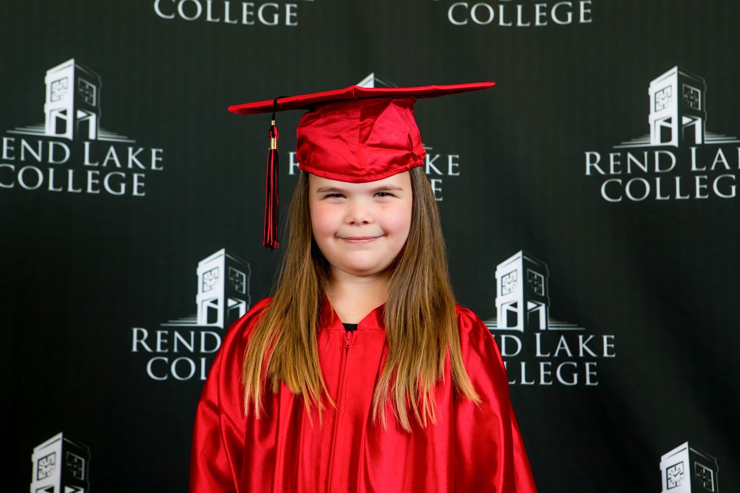 A studio portrait of a young girl with long, straight brown hair offering a pleasant smile. She wears a vibrant red graduation gown and a matching red cap with a hanging tassel. The backdrop is solid black with the white "Rend Lake College" wordmark and clock tower icon repeated uniformly.