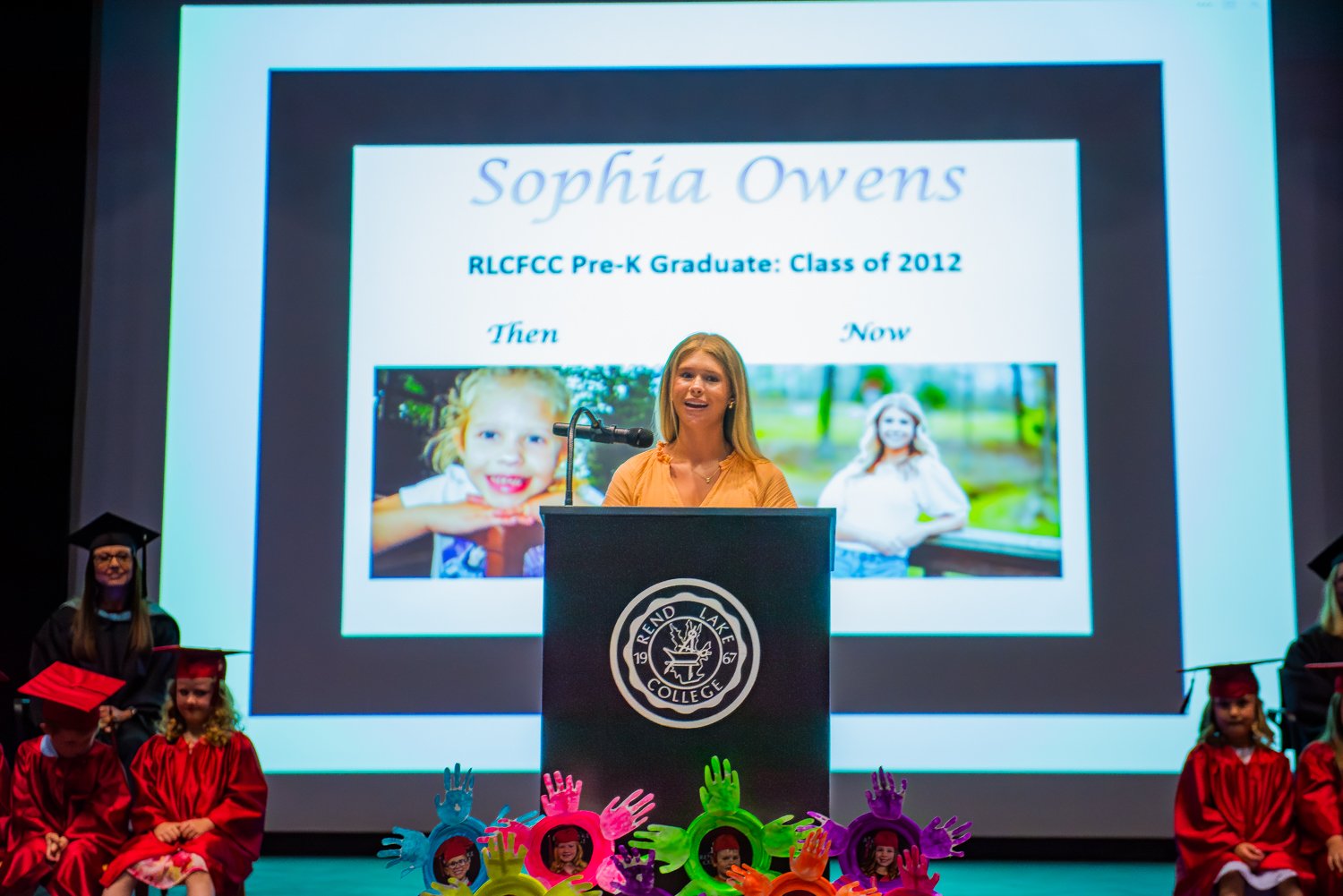 A young woman with long blonde hair, Sophia Owens, speaks at a microphone behind a dark podium bearing the circular Rend Lake College seal. Behind her, a large projection screen displays a slide titled "Sophia Owens / RLCFCC Pre-K Graduate: Class of 2012." The slide features a side-by-side comparison labeled "Then" (a childhood photo of her smiling as a young girl) and "Now" (a recent portrait of her as a high school senior). At the base of the podium, colorful, handprint-shaped picture frames holding photos of young graduates are lined up. In the background, seated on either side of the stage, a teacher in a black academic gown and several young Pre-K graduates in shiny red caps and gowns watch her speak.