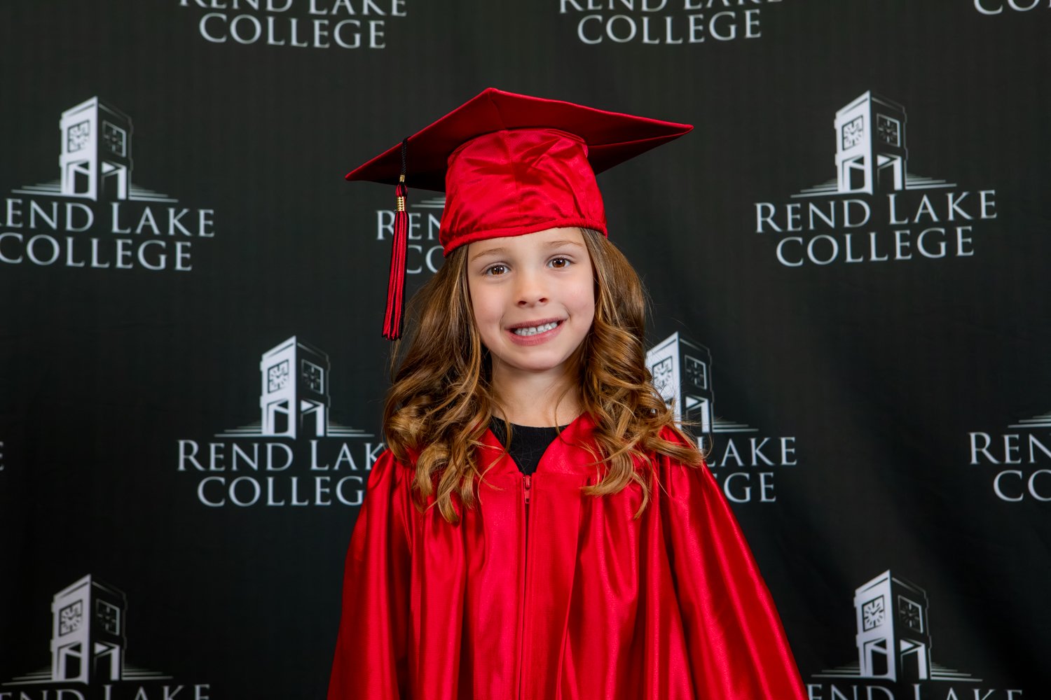 A studio portrait of a young girl with long, curling brown hair smiling broadly. She is dressed in a glossy red graduation gown and a matching red mortarboard cap. She is framed against a black background featuring a repeating white pattern of the "Rend Lake College" institutional logo.