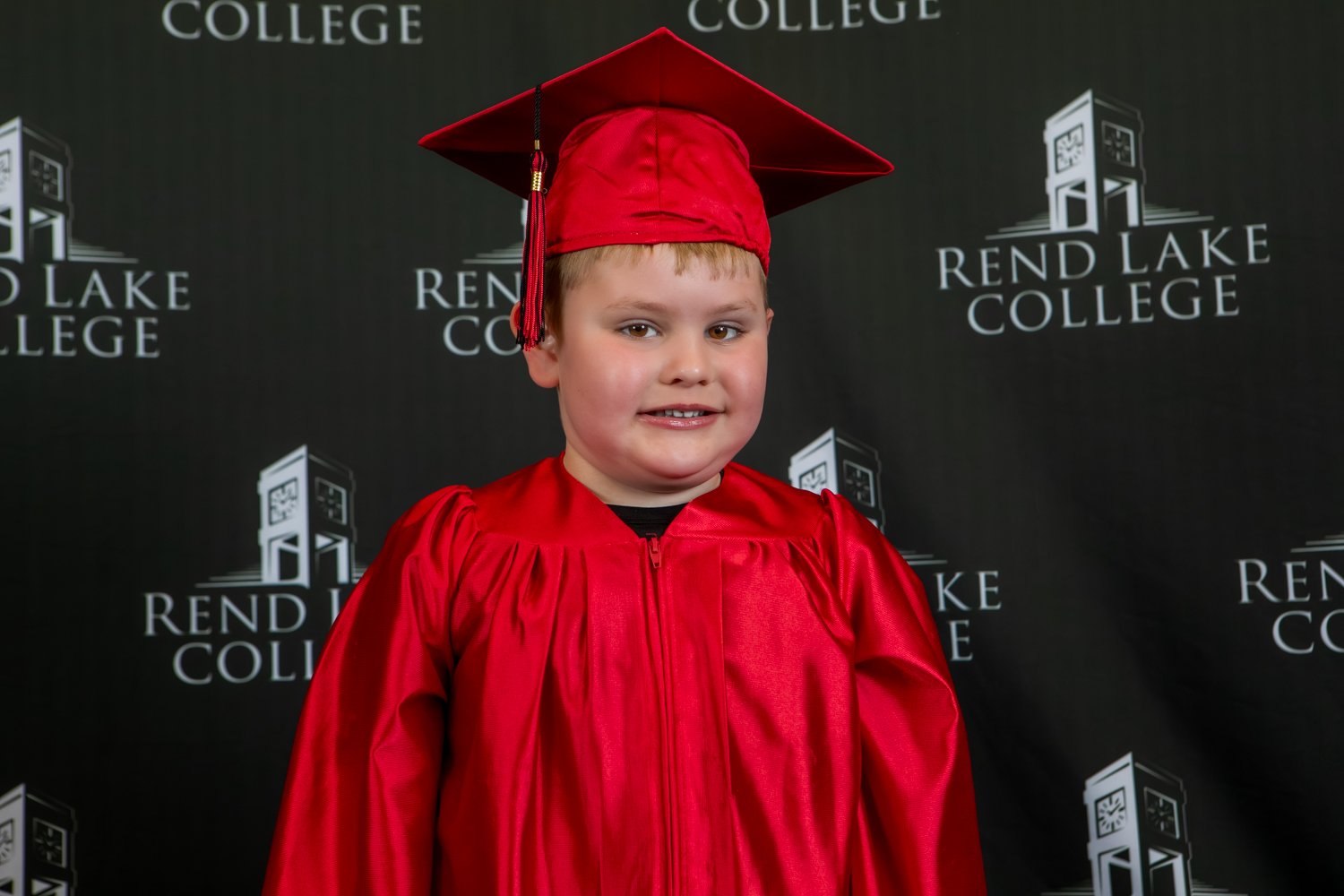 A studio portrait of a young boy with short brown hair smiling warmly. He is wearing a shiny red graduation gown and a matching red mortarboard cap with a dark tassel hanging to his right. He stands centered against a solid black backdrop repeating the "Rend Lake College" logo and clock tower icon in white.