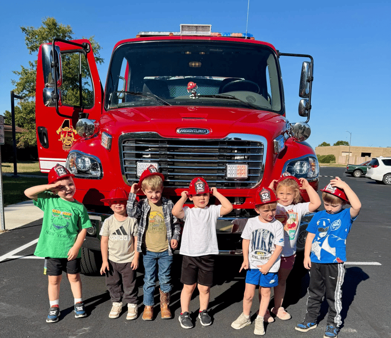 Seven young children stand in a row in a paved parking lot in front of a large, red Freightliner fire truck on a sunny day. They are wearing red toy firefighter hats; one boy on the left salutes, while others hold onto their hats or pose for the camera.