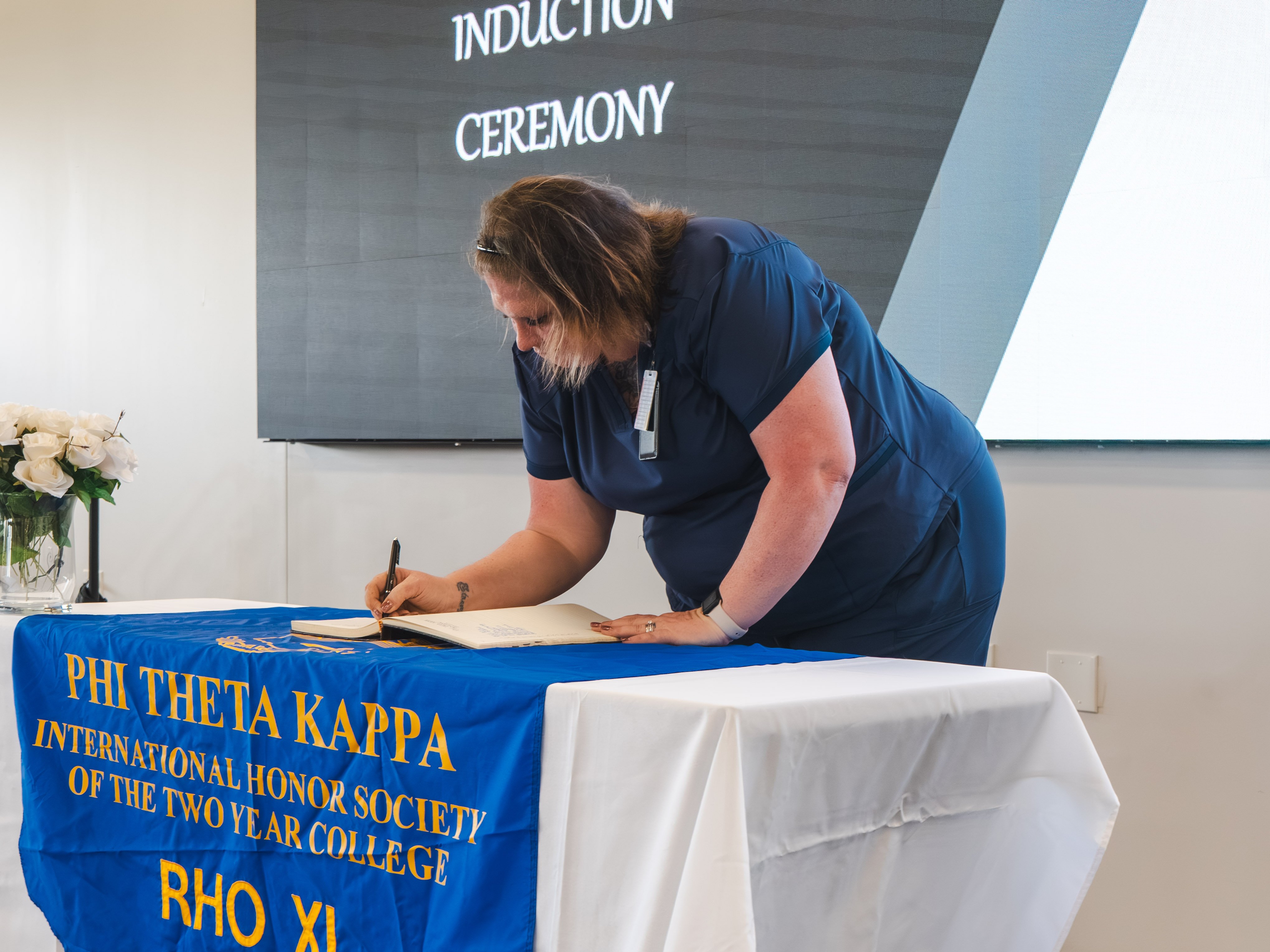 A female inductee dressed in navy blue medical scrubs leans over a table to sign the induction book. She has shoulder-length brown hair and an ID badge clipped to her collar. The table features a blue banner with gold text reading "Phi Theta Kappa International Honor Society of the Two Year College."