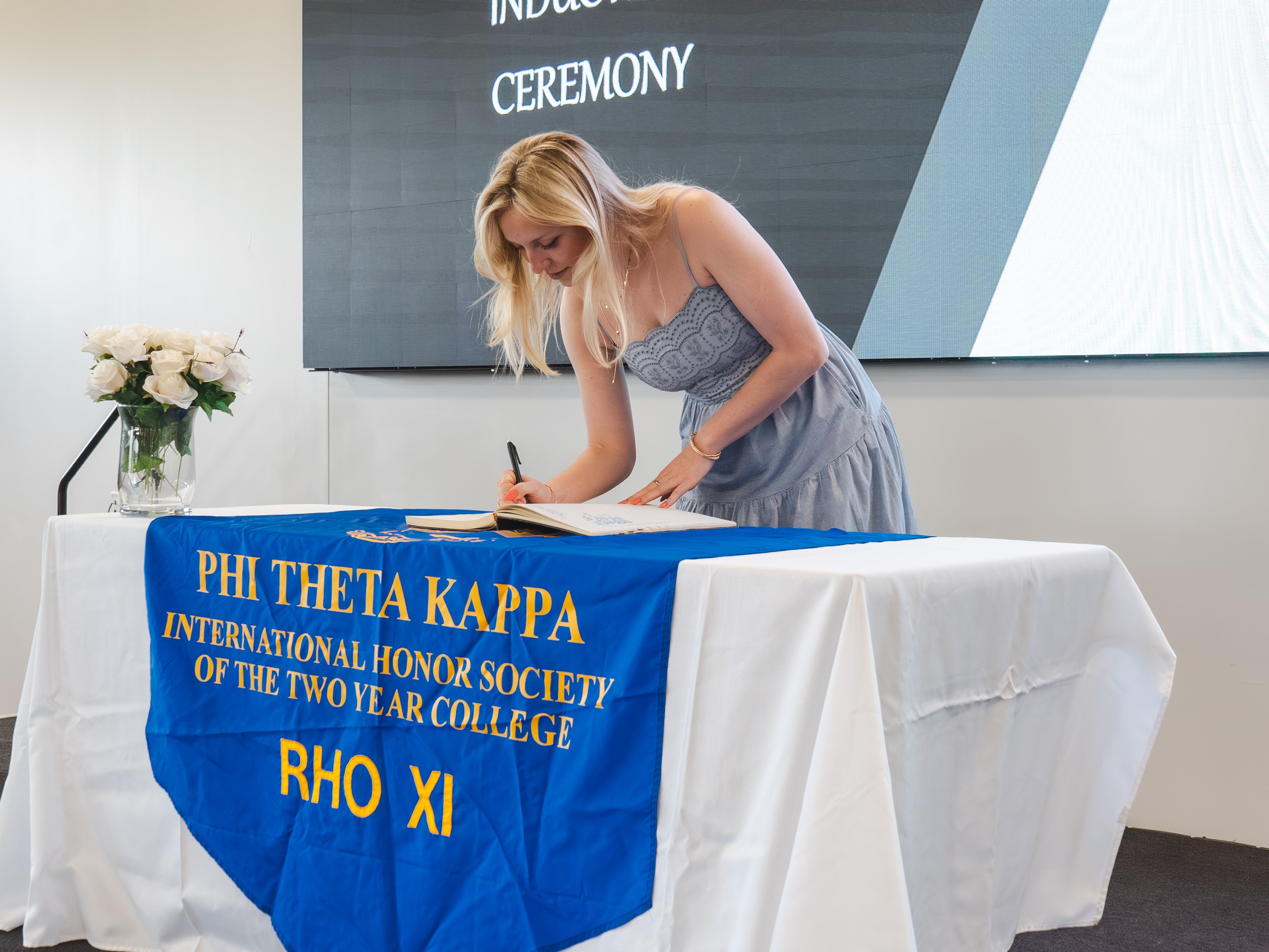 A female inductee with blonde hair leans over a white-clothed table to sign a guest book. She is wearing a light blue, patterned sundress with spaghetti straps and a gold bracelet. The table is adorned with a blue Phi Theta Kappa banner. A large screen displaying "Induction Ceremony" sits in the background.