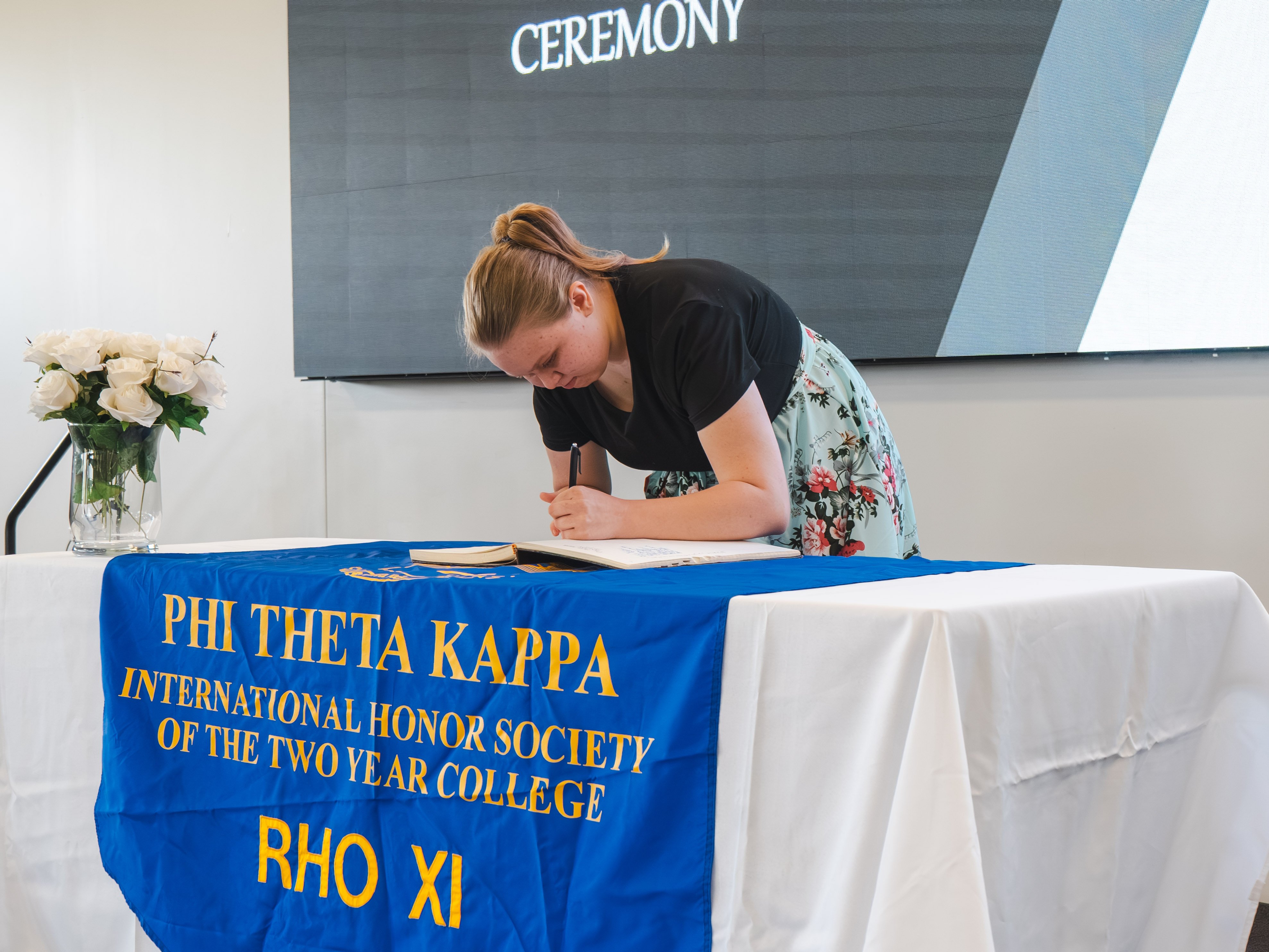 A female inductee with her blonde hair pulled back into a ponytail leans forward to sign the induction log. She is wearing a black t-shirt and a light mint-green skirt with a red and green floral pattern. She stands at a white registration table featuring a blue and gold Phi Theta Kappa banner.