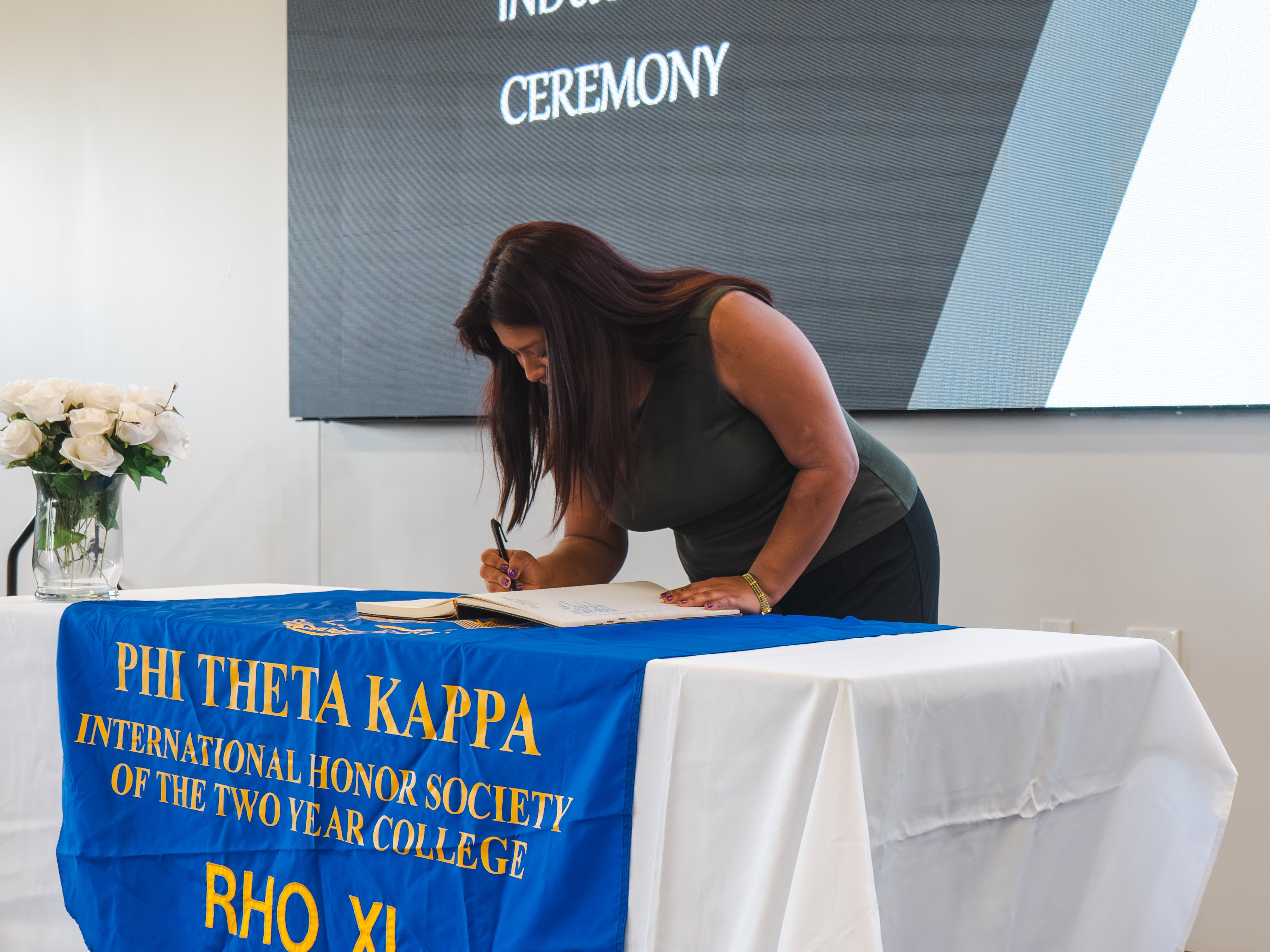 A female inductee with long, dark reddish-brown hair leans over a table to sign the official book. She is wearing a sleeveless olive-green top and black slacks. The table is decorated with a white tablecloth and a blue Phi Theta Kappa honor society banner.