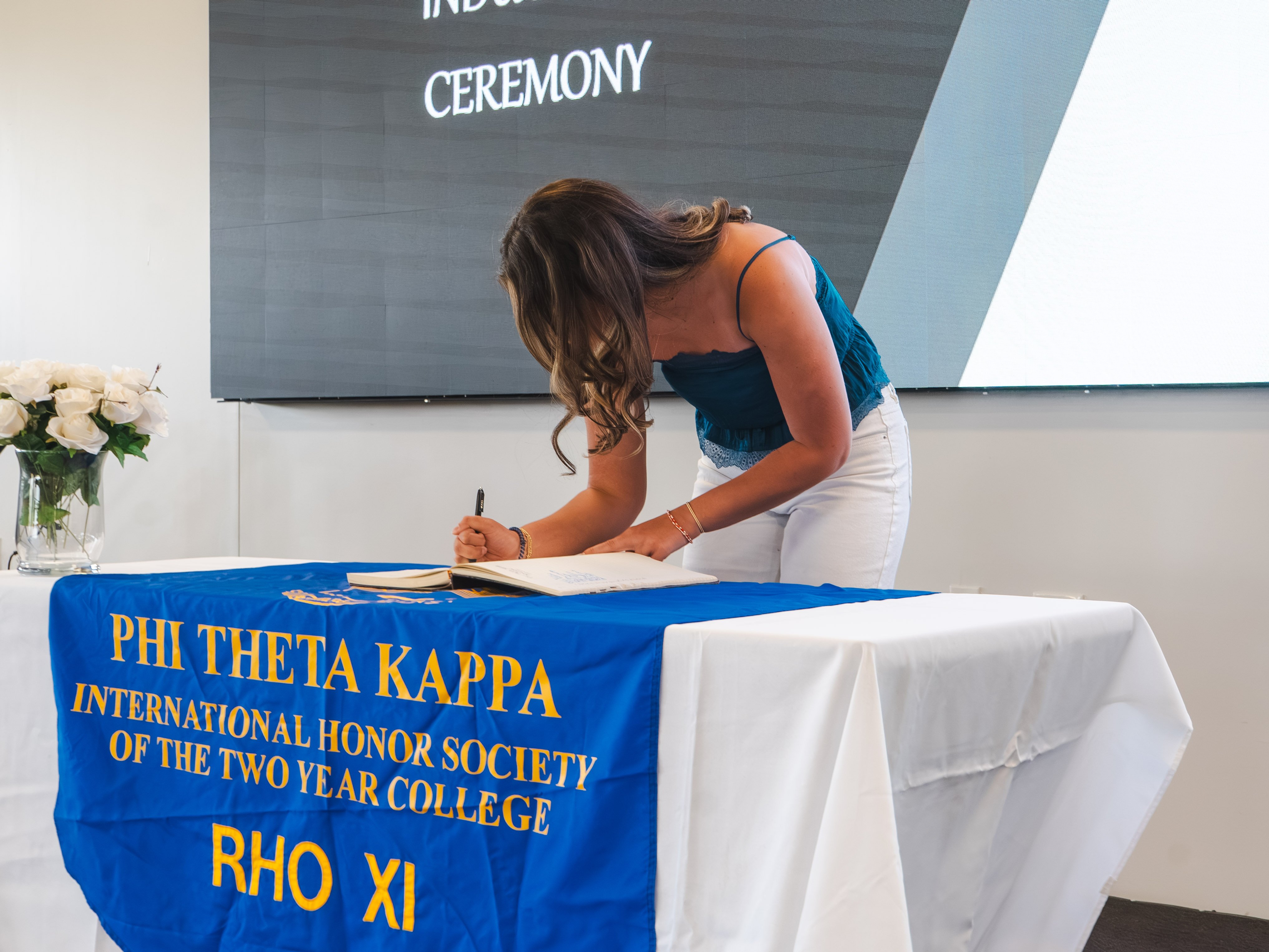 A female inductee with long, wavy brown hair leans down to sign the registry. She is wearing a teal blue tank top and white pants. She stands at a table draped in a blue and gold Phi Theta Kappa banner, with a digital event screen visible in the background.