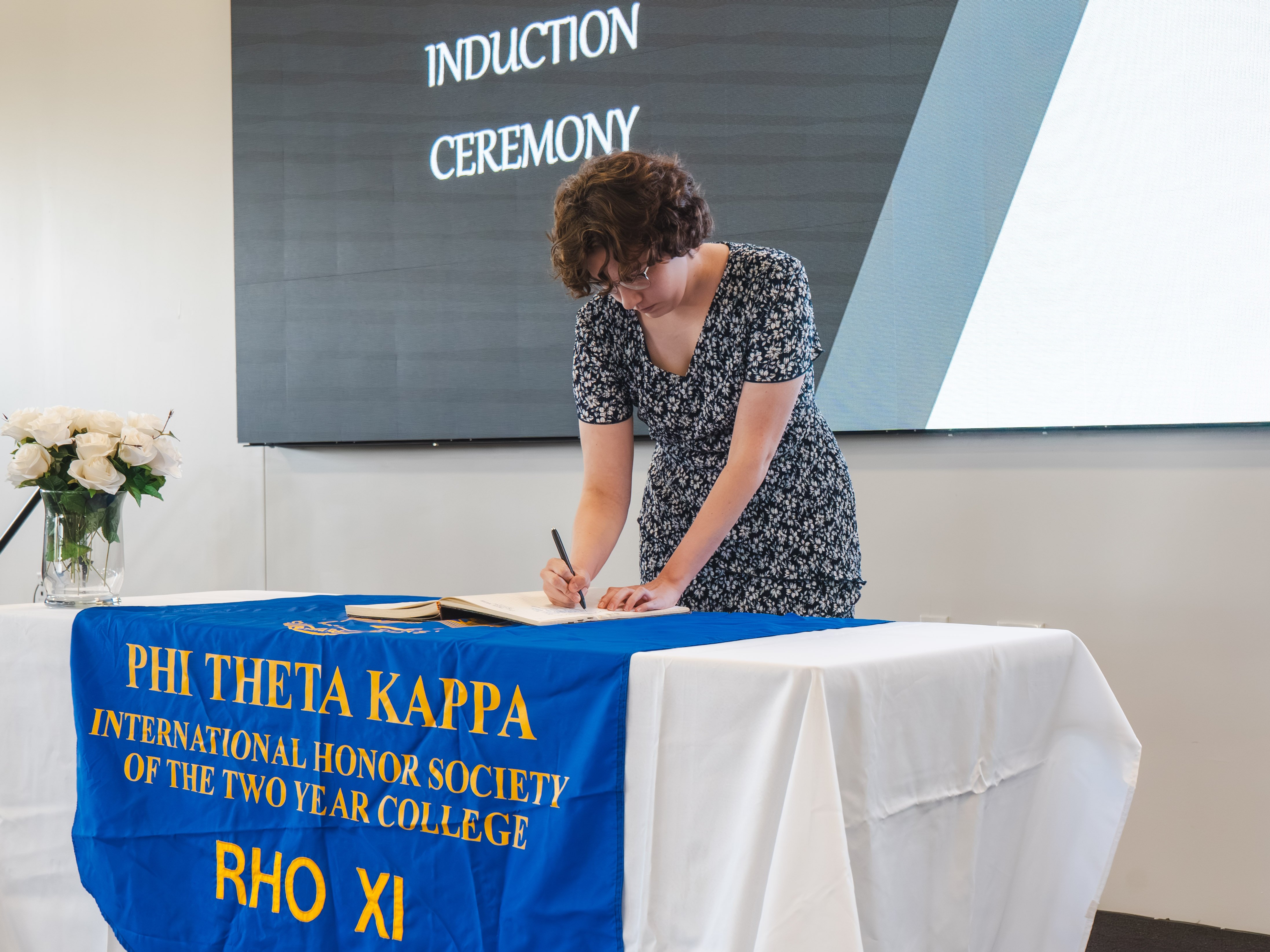 A female inductee with short, curly brown hair and glasses leans forward to sign the induction book. She is wearing a dark blue and white patterned V-neck dress. The table is covered with a blue banner displaying the gold text "Phi Theta Kappa International Honor Society of the Two Year College."