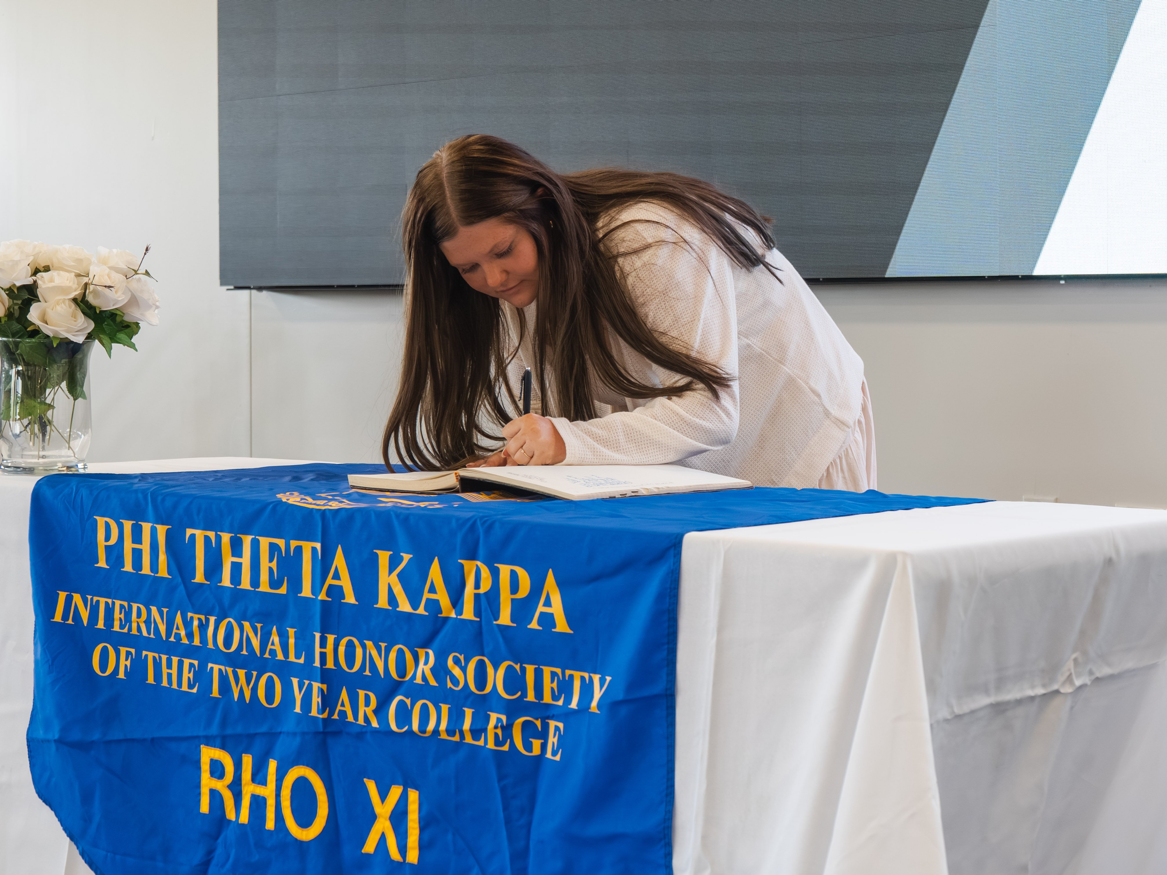 A female inductee with long, straight dark hair leans over a white-clothed table to sign the registration book. She is wearing a cream-colored, textured long-sleeve blouse. The table is draped with a bright blue Phi Theta Kappa honor society banner displaying gold text. A vase of white roses sits to her left against a neutral background.