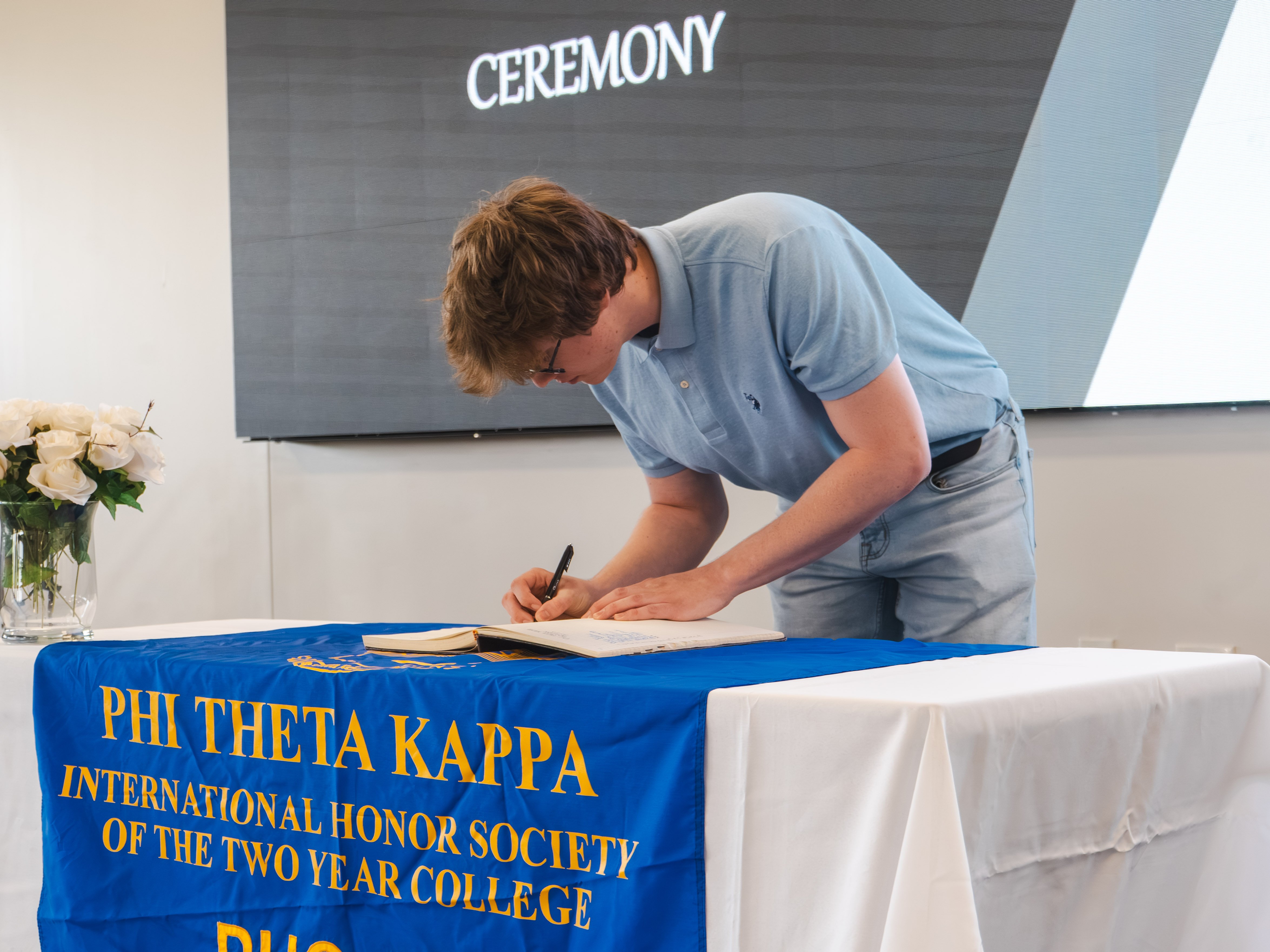 A young male inductee with wavy light brown hair and glasses leans down to sign his name in the official induction book. He wears a light blue polo shirt and light-wash jeans. He is standing at a presentation table adorned with a blue and gold Phi Theta Kappa banner and a bouquet of white roses.