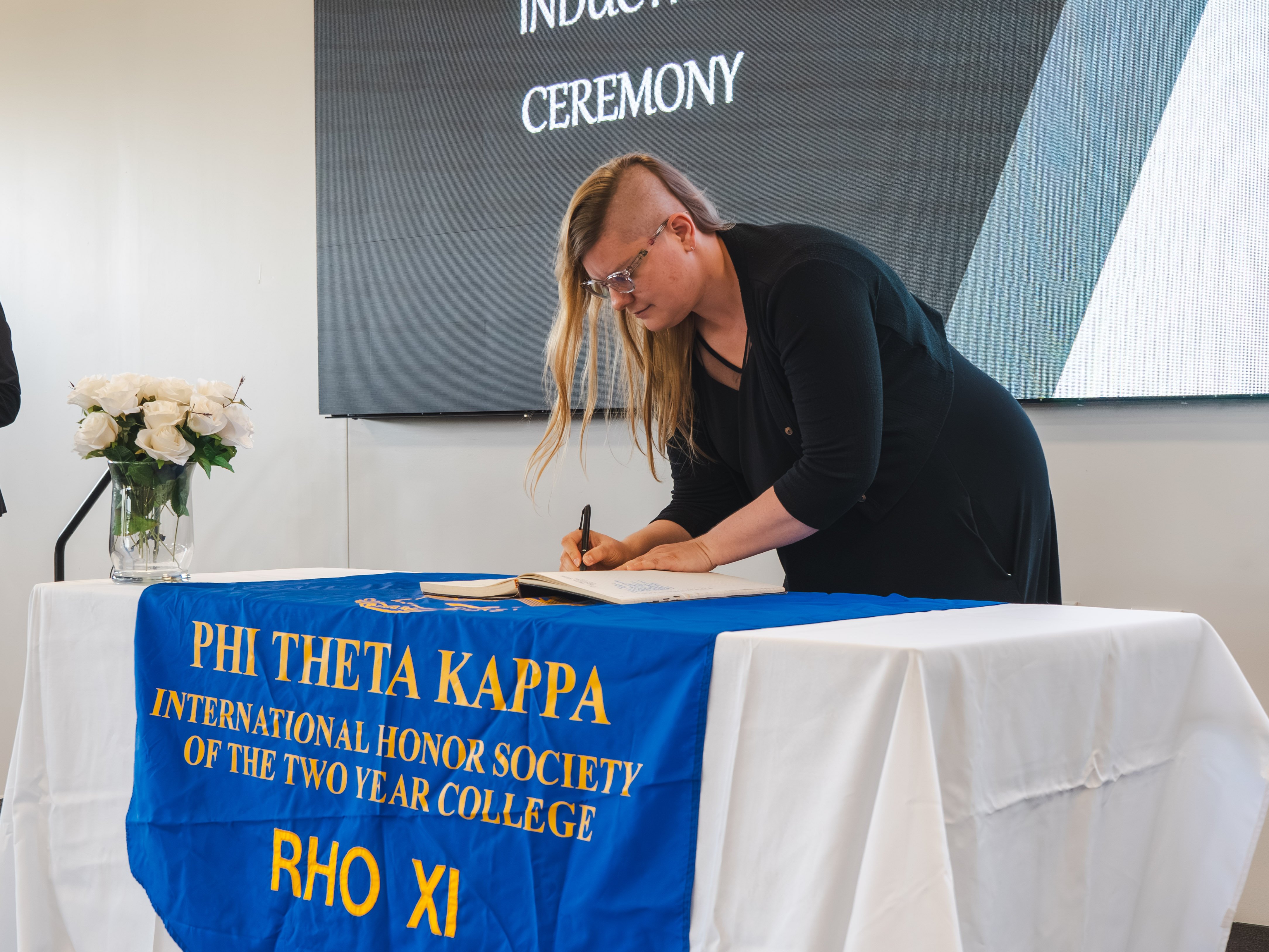A female inductee with blonde hair styled with a side-shave haircut and wearing glasses leans over the table to sign the ledger. She is dressed in a black top and black cardigan. The table features a blue Phi Theta Kappa banner with gold lettering. A large digital screen sits in the background.