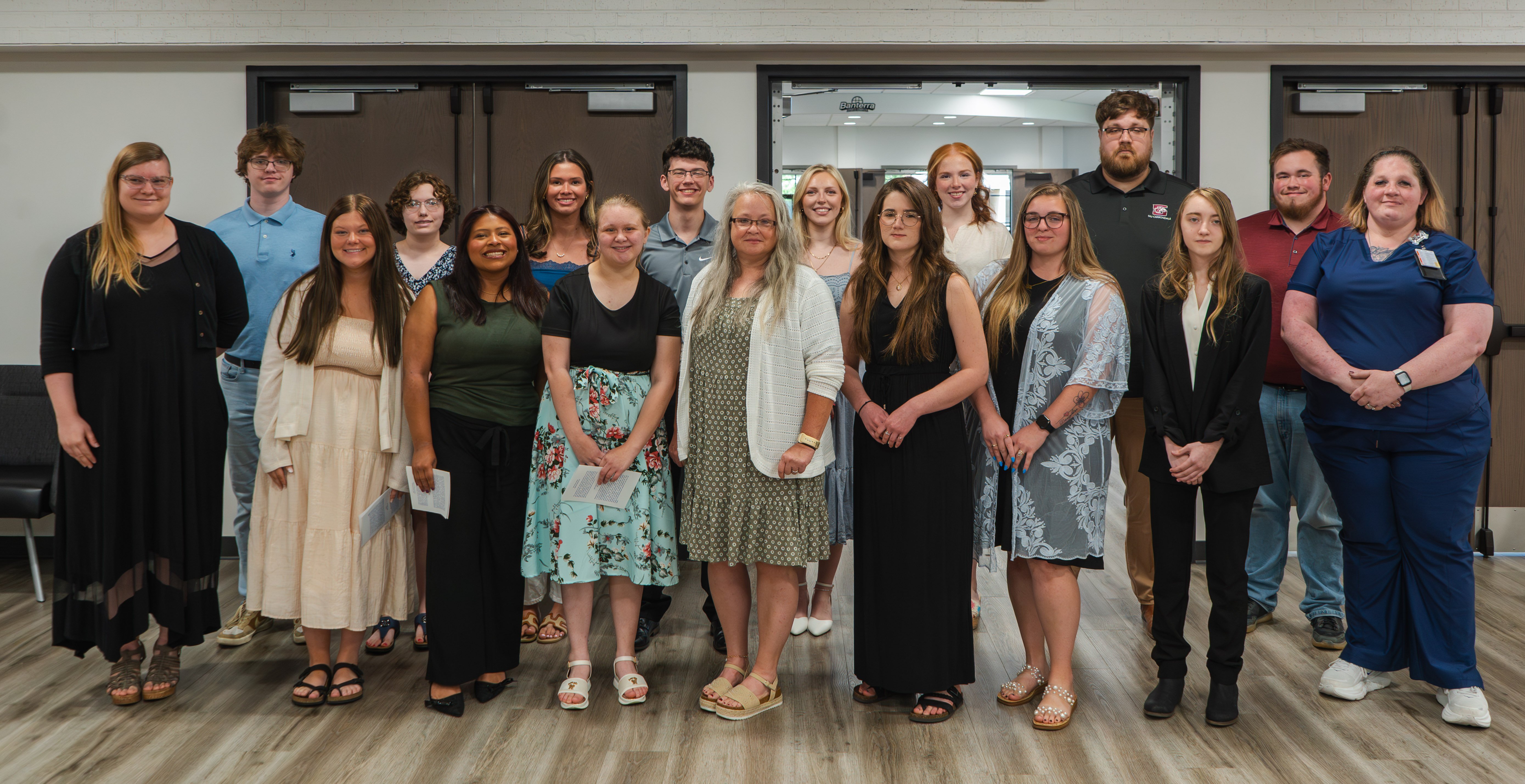 A group of fifteen individuals stand together indoors for a formal group photograph following a Phi Theta Kappa induction ceremony. The group is arranged in two rows, with most members standing side-by-side facing the camera. They are dressed in a mix of professional and smart-casual attire, including dresses, blouses, button-down shirts, and slacks. One individual on the far right wears navy blue medical scrubs. Several people in the front row hold small, folded program papers.