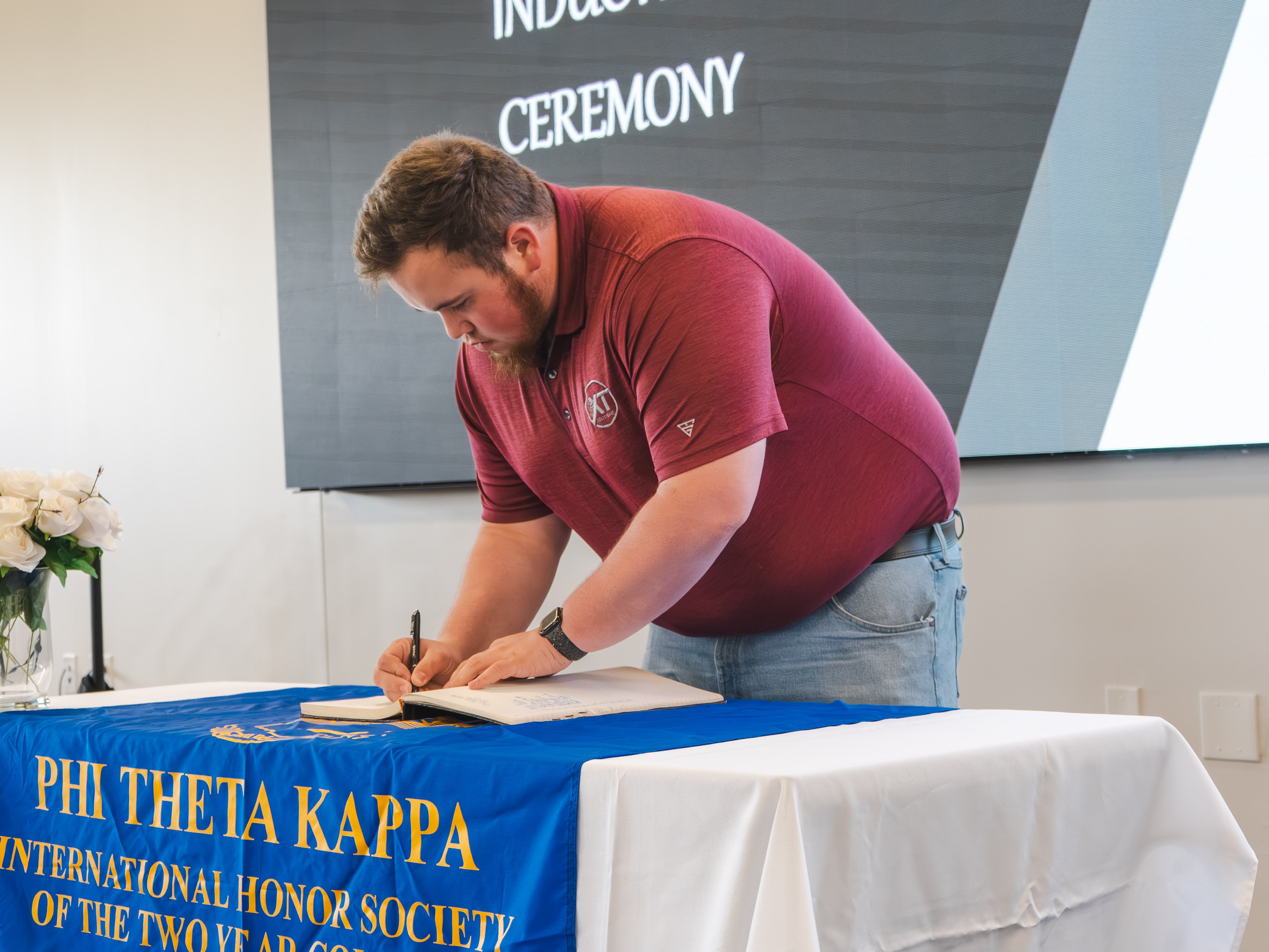 A male inductee with short brown hair and a beard leans over a table to sign the registry. He is wearing a maroon polo shirt and light-wash blue jeans. The table is decorated with a white tablecloth and a blue Phi Theta Kappa honor society banner.