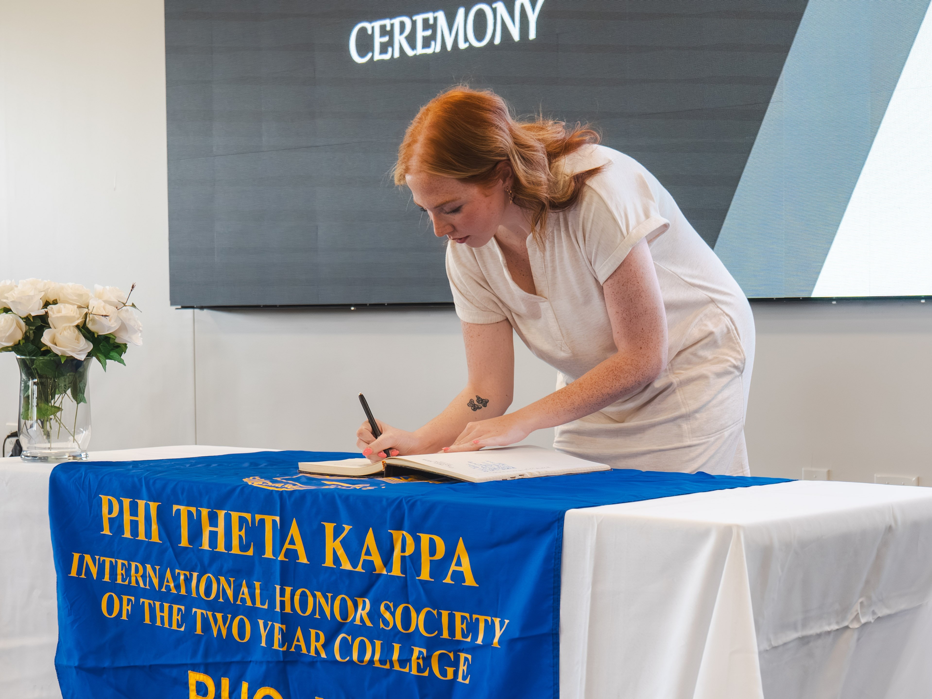 A female inductee with red hair pulled back leans down to sign the guest book during the ceremony. She wears a short-sleeved white dress, revealing a small butterfly tattoo on her inner forearm. She stands at a table with a blue and gold Phi Theta Kappa banner.