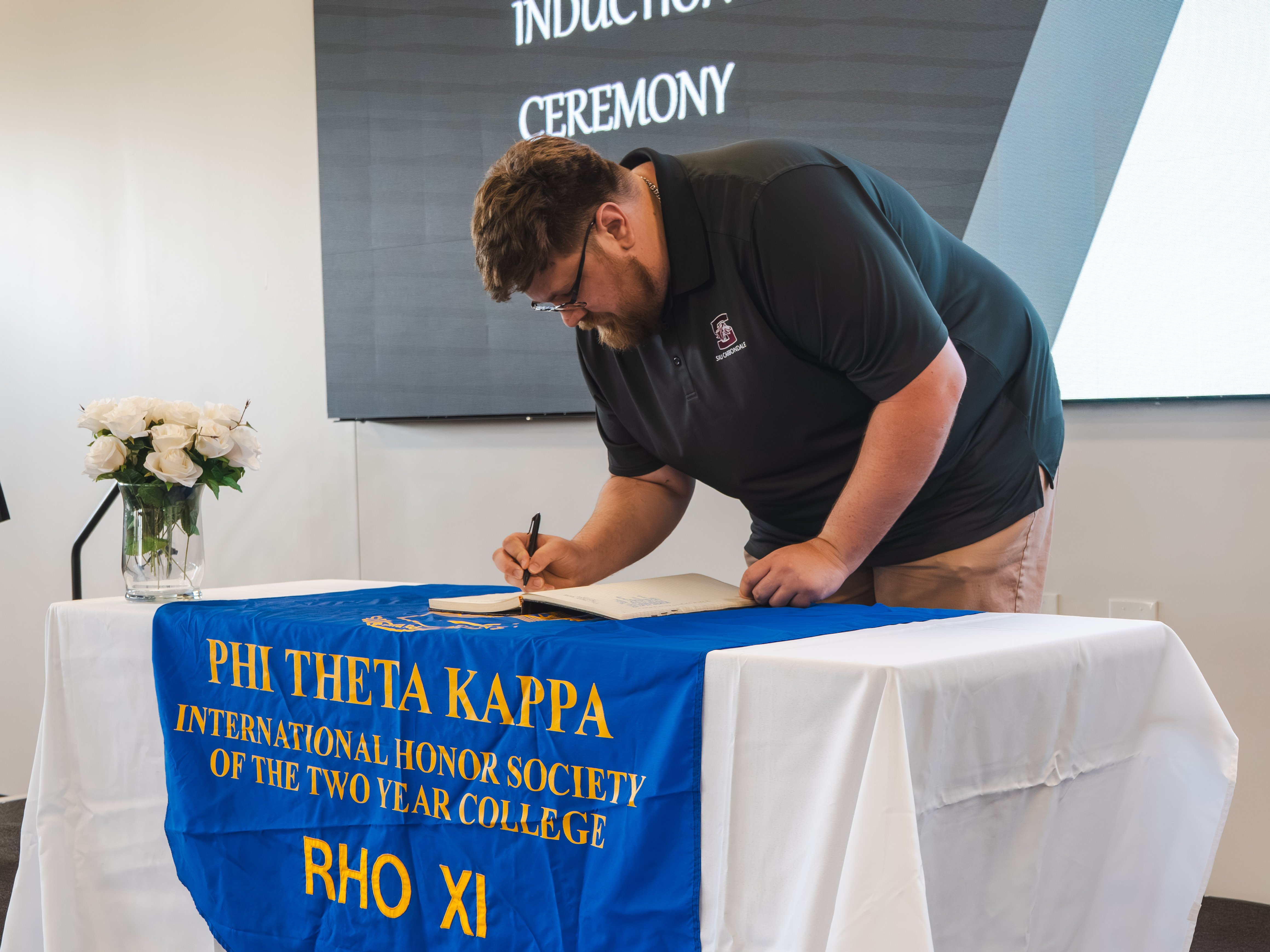 A large-statured male inductee with dark hair, glasses, and a beard leans over the table to sign the induction book. He is wearing a dark grey polo shirt with a small collegiate logo and khaki pants. The table is covered with a blue Phi Theta Kappa banner.