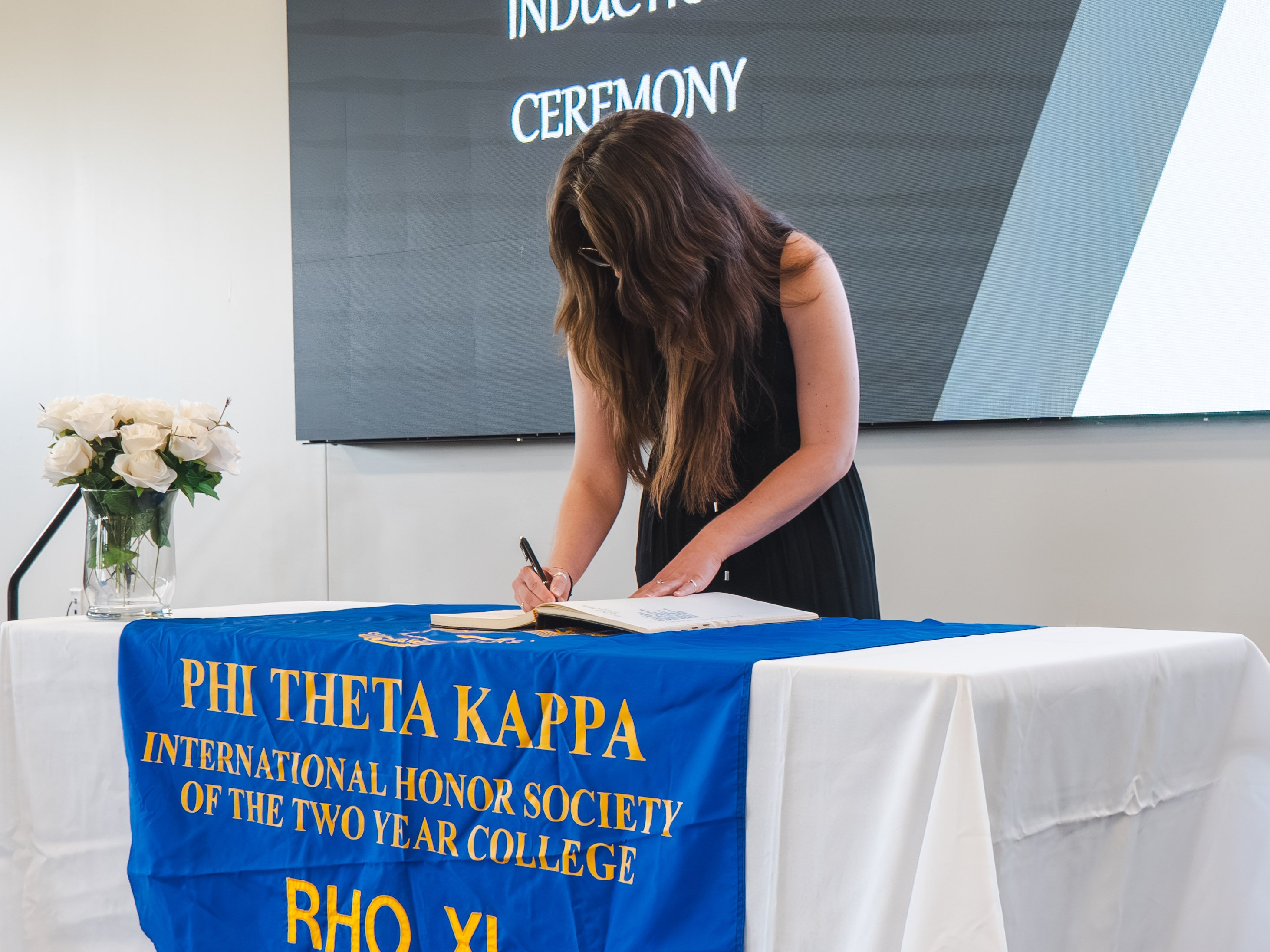 A side-angle view of a female inductee with long, dark brown hair leaning over a table to sign an induction book during a Phi Theta Kappa ceremony. She wears a sleeveless black dress and glasses. The table is draped in a white tablecloth and covered with a vibrant blue banner featuring gold lettering that reads "Phi Theta Kappa International Honor Society of the Two Year College." A glass vase containing white roses sits on the left side of the table, and a large digital screen displaying "Induction Ceremony" is visible in the background.