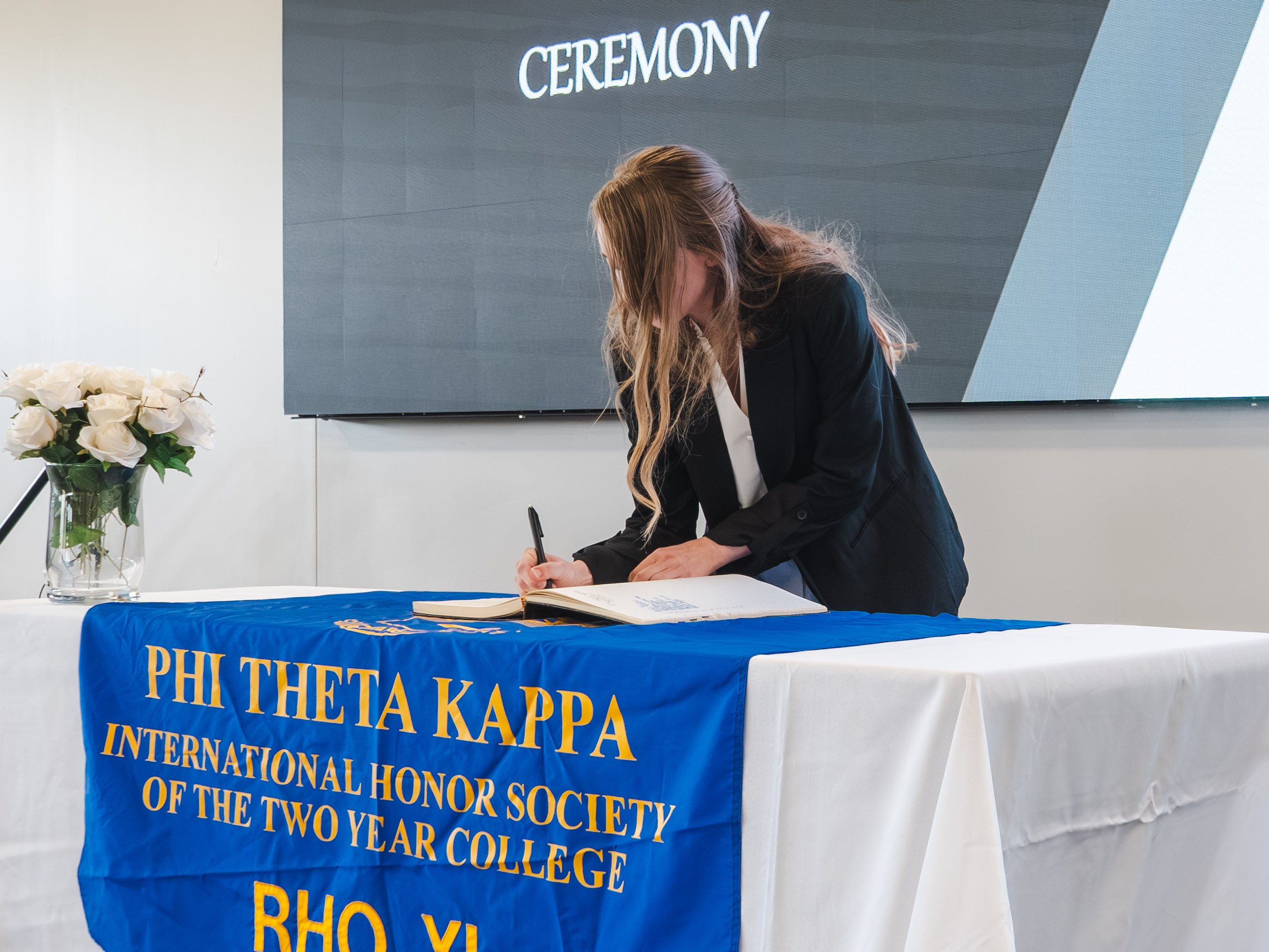 A medium shot of a female inductee with blonde hair pulled back in a half-up style, leaning forward to sign the official registry book. She is wearing a professional black blazer over a white blouse. She stands at a white-clothed table decorated with a blue and gold Phi Theta Kappa banner and a vase of white roses. The background features a large presentation screen displaying the word "Ceremony."