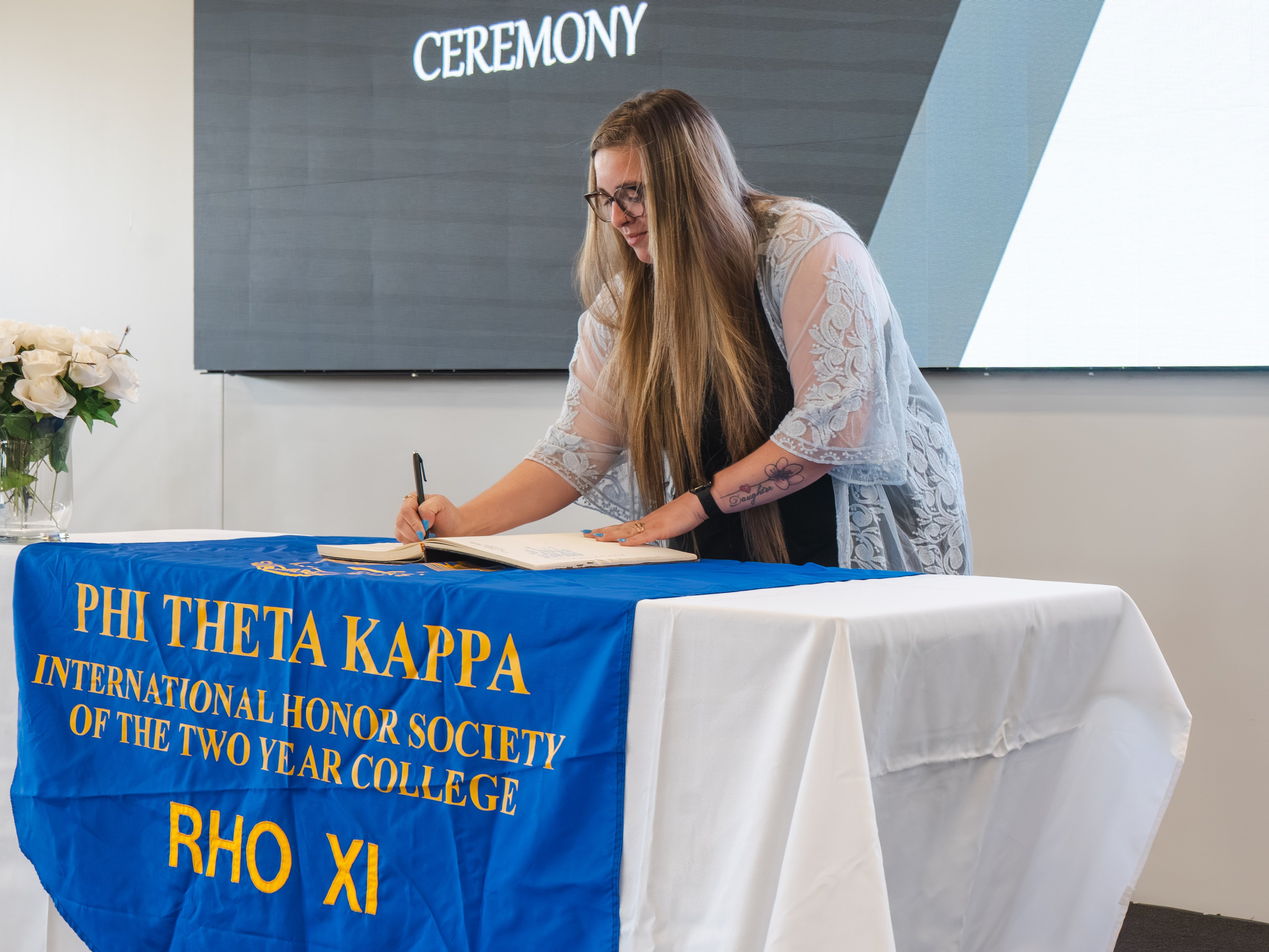 A female inductee with long straight hair and glasses leans over a registration table to sign the induction logbook. She wears a black top layered with a light blue, sheer, lace-detailed cardigan that shows a small wrist tattoo. The table features a blue and gold Phi Theta Kappa honor society banner. A large digital screen and a bouquet of white roses are in the background.