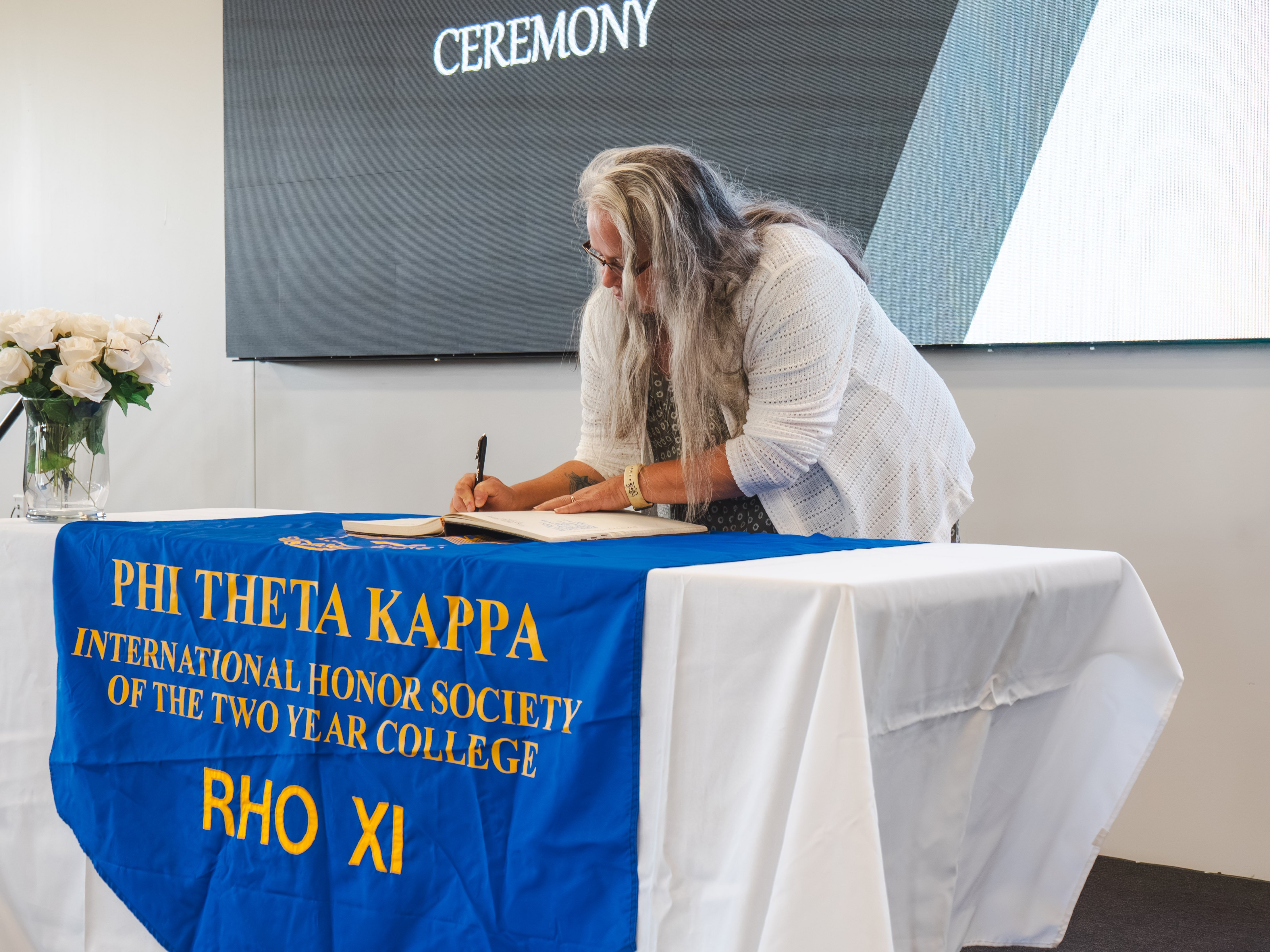 An inductee with long, wavy grey and silver hair leans down to sign the induction book. She is wearing a dark, patterned dress and an open-knit white cardigan. The table is covered with a white cloth and a blue Phi Theta Kappa chapter banner. A large presentation screen is visible behind her.