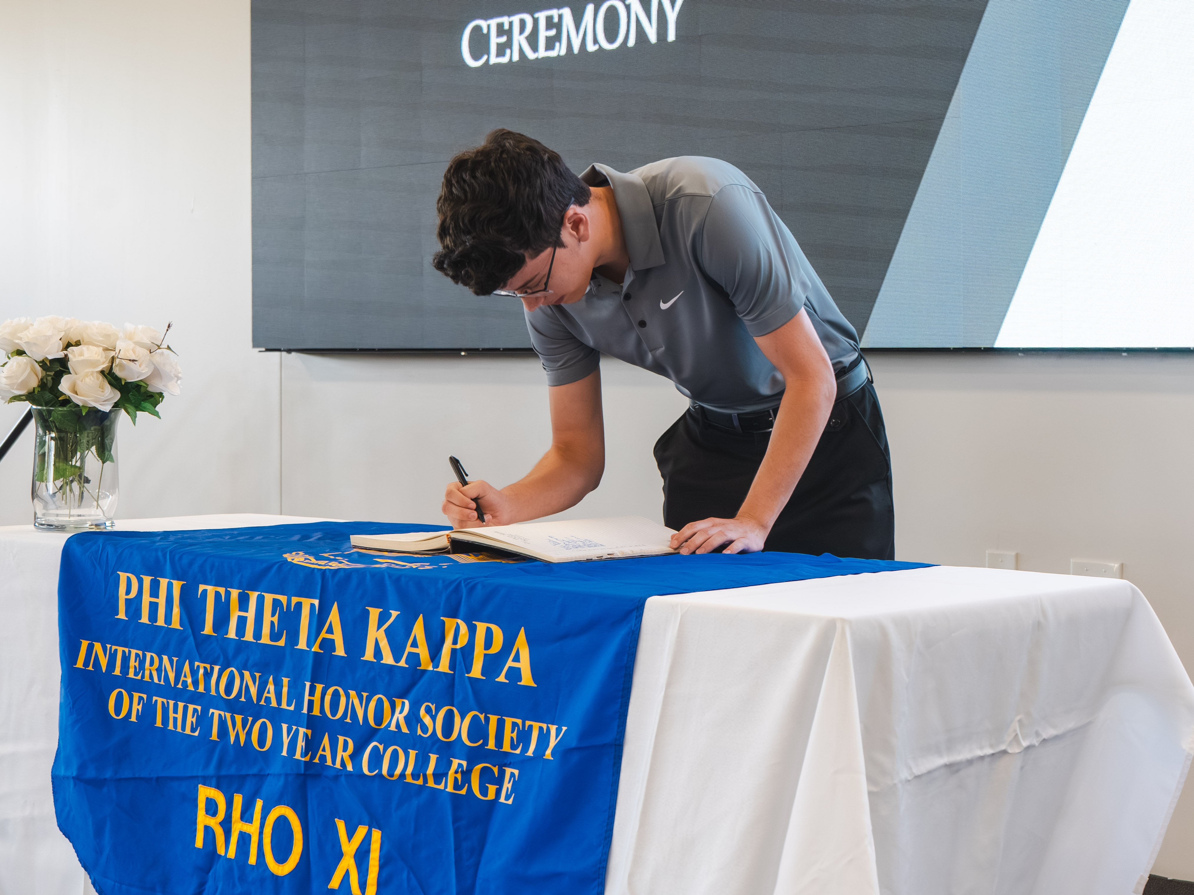 A young male inductee wearing glasses and a grey Nike polo shirt leans deeply over a table to sign his name in the official ledger. He is positioned at a table draped with a white cloth and a blue and gold Phi Theta Kappa banner. The background shows a digital event screen and a vase of white roses.