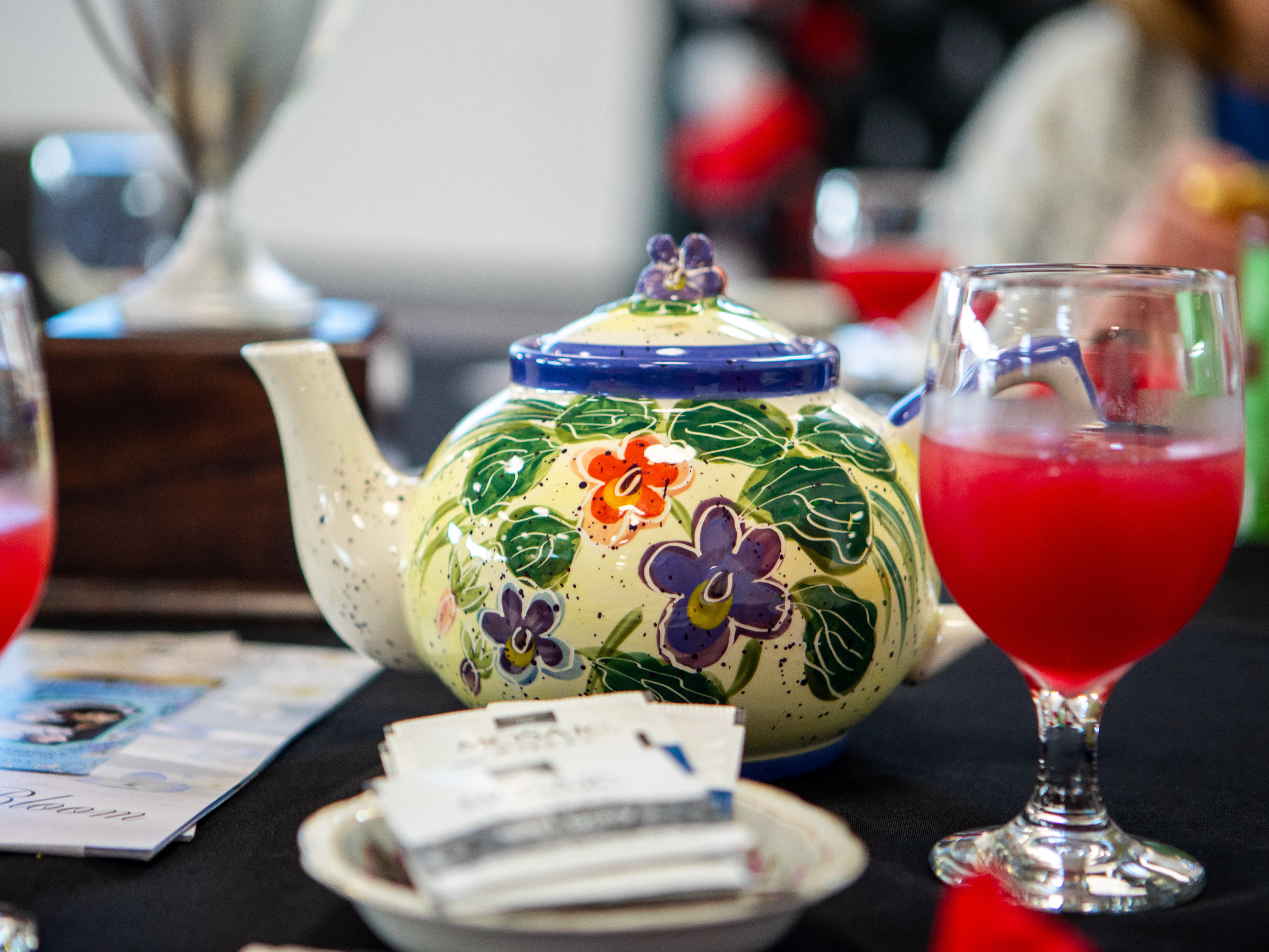 A close-up of a table setting featuring a decorative, cream-colored teapot painted with green leaves and purple and orange flowers. Next to the teapot sits a stemmed glass filled with a bright red beverage and a small dish holding several tea bags, all resting on a black tablecloth.