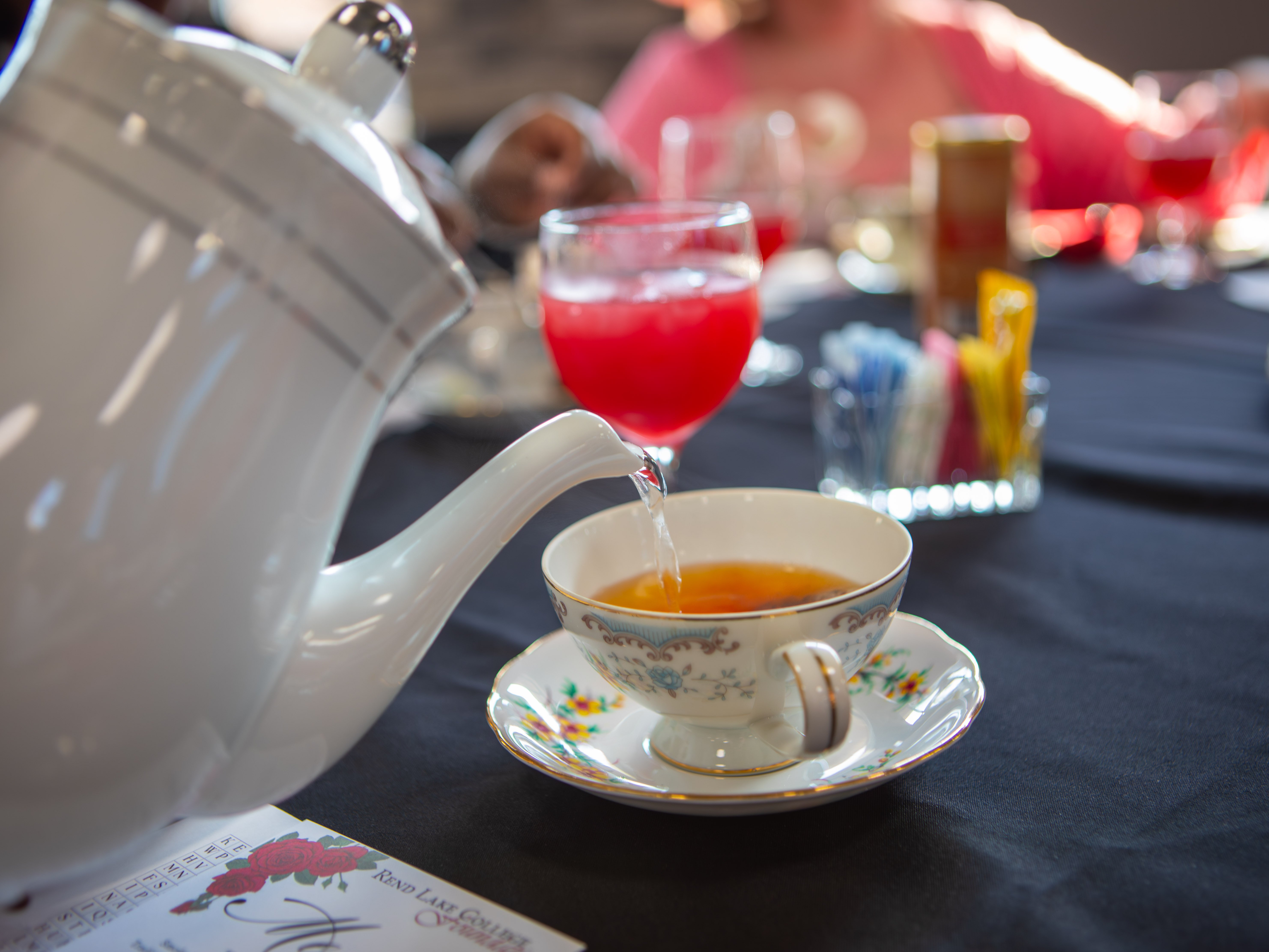 A white ceramic teapot pours steaming tea into an elegant teacup on a matching saucer decorated with delicate blue and yellow flowers.
