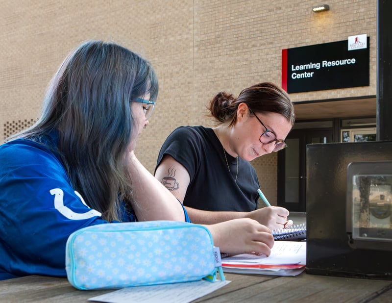 Two students sit together at an outdoor table studying and writing in notebooks outside the Rend Lake College Learning Resource Center.