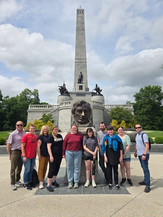 A group of ten TRiO STARS students and staff pose outdoors in front of Lincoln's Tomb in Springfield, Illinois, standing before the large bronze bust of Abraham Lincoln and the tall obelisk monument under a partially cloudy sky.