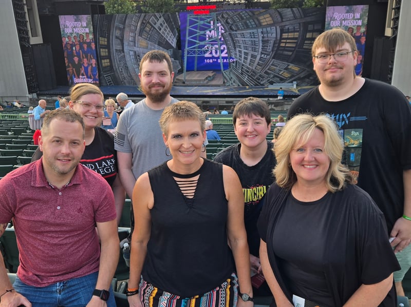 A group of six TRiO STARS students and staff smile together for a photo in the outdoor seating area at The Muny theater in St. Louis, with the stage lit up in the background before a performance.