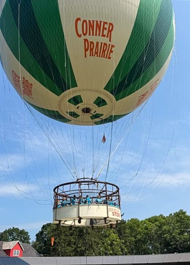 A massive green and beige hot air balloon emblazoned with the words "Conner Prairie" floats high in a clear blue sky. A large, circular gondola carrying a group of Upward Bound students in blue shirts hangs suspended below the balloon as it hovers over lush green trees and a red building.