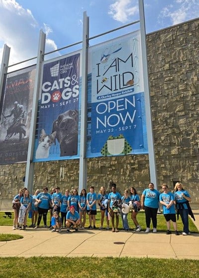 A large group of Upward Bound students and leaders wearing matching blue t-shirts line up outside a museum building. Large outdoor banners hang behind them advertising IMAX, a "Cats & Dogs" exhibit, and the "I Am Wild" Charley Harper exhibit at the Indiana State Museum.