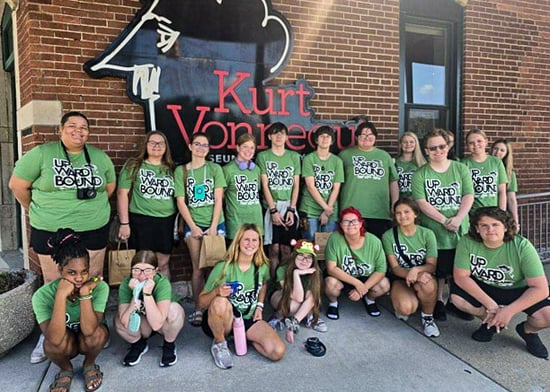 A group of Upward Bound students and staff, all wearing matching green t-shirts, pose for a photo together outside the brick facade of the Kurt Vonnegut Museum. Some group members are kneeling in the front row while others stand behind them in front of the museum's black and red logo sign.