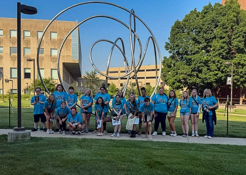 A large group of Upward Bound students and staff, all wearing matching light blue t-shirts, pose together on a concrete sidewalk on the Purdue University campus. Behind them stands a tall, abstract metal loop sculpture set against green campus lawns, trees, and university buildings.
