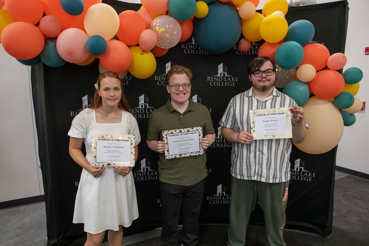 Three high school students stand side-by-side in front of a black backdrop with the Rend Lake College logo, adorned with a large arch of vibrant orange, yellow, pink, and teal balloons. From left to right, Hailey Tomshack wears a white short-sleeved dress and holds her certificate with a neutral expression; Emerson Vincelette wears a green polo shirt, glasses, and smiles brightly while holding his award; Wyatt Shively wears a striped, short-sleeved button-down shirt, glasses, and green sweatpants, holding his certificate with a slight smile. Each holds a "Certificate of Achievement" for perfect attendance in the Upward Bound Summer Program.