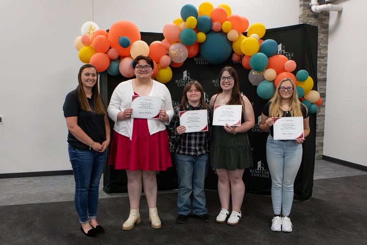 A group of five women stand in a line posing for a photo in front of a black backdrop with the Rend Lake College logo, which is topped by a large arch of orange, yellow, and teal balloons. From left to right, RLC Foundation Assistant Director Jessica Jackanicz stands smiling in a black polo and jeans. Next to her, four students stand smiling and holding their scholarship certificates: Lydia Robison in a red dress and white cardigan, Rylee Tramee in a black and white plaid shirt and jeans, Shauna Wilkerson in a black patterned sundress, and Kira Crane in a black top and light-wash jeans.