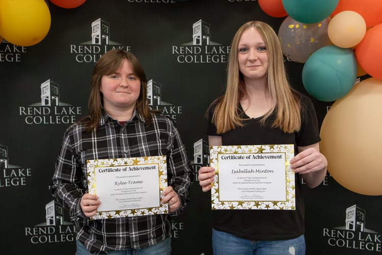 Two high school students stand side-by-side holding "Certificate of Achievement" awards in front of a black backdrop patterned with the Rend Lake College logo. On the left, Rylee Trame wears a black and white plaid button-down shirt and smiles gently while holding her certificate. On the right, Isabellah Minton wears a black t-shirt and blue jeans, holding her certificate with a smile. The awards recognize their straight A's in the Upward Bound Summer Program. The scene is decorated with festive orange, yellow, and teal balloons framed at the top and right sides.