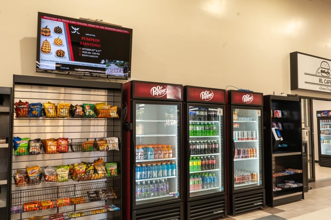 A convenience-style snack and beverage station inside a building with beige walls and tiled flooring. On the left, a wire rack shelf holds a variety of bagged chips and boxed snacks. Above the rack hangs a wall-mounted digital display showing an announcement for a "Pumpkin Painting" event at Rend Lake College.   In the center stand three glass-door upright coolers branded with the Dr Pepper logo at the top, filled with bottled and canned drinks including Gatorade, sodas, and juices. To the right of the coolers is a dark wood shelving unit with small impulse items and payment kiosk equipment, followed by a glass doorway labeled "RL Cafe".  