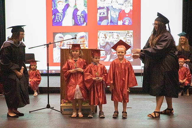 Three young children wearing red caps and gowns stand on stage during the 2022 Rend Lake College Foundation Children's Center Pre-K graduation ceremony while staff members assist them.