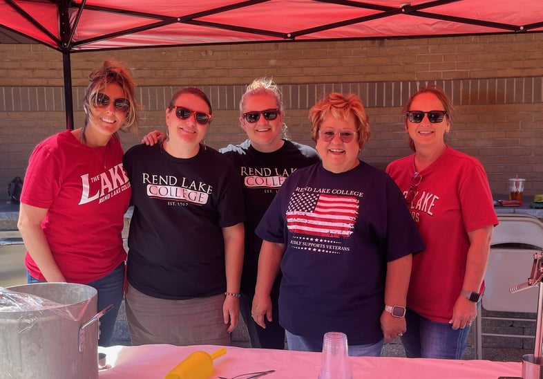 Five smiling RLC-Cares team members in sunglasses and college t-shirts under a red tent.