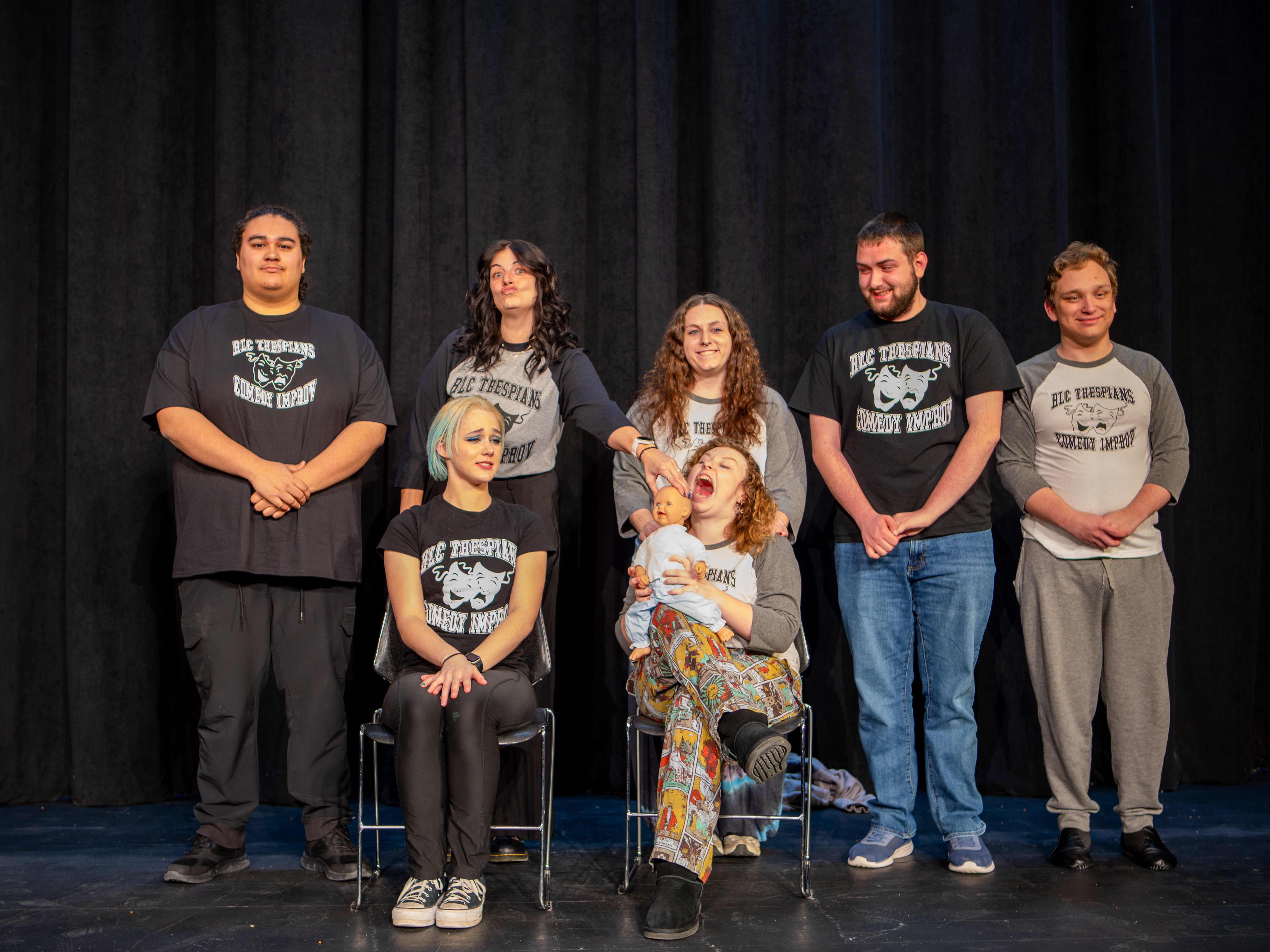 Seven members of the RLC Thespians Comedy Improv team pose on a stage in front of a black curtain, wearing matching shirts featuring drama masks. In the front row, Madison Brower (left) has blue hair and a somber expression, while Jinx Craddock (right) holds a baby doll and laughs with their mouth wide open. In the back row, standing from left to right, are Malcom Hall, Taylor Wilburn (making a playful face), Gabrielle Webb, Jack Robison, and Samuel Buesking.